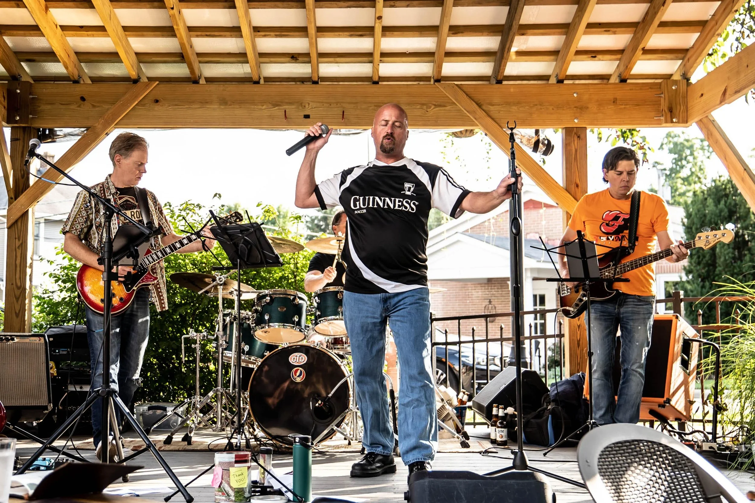 A band performing on an outdoor stage with three musicians playing guitars and drums, one singer in front holding a microphone, under a wooden roof, with trees and houses in the background.