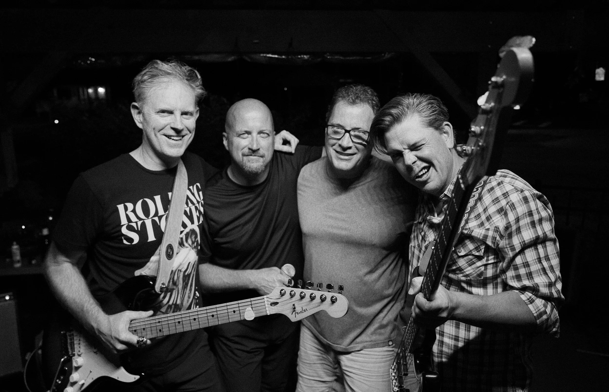 Four men with musical instruments, smiling and posing together in a dark indoor setting.