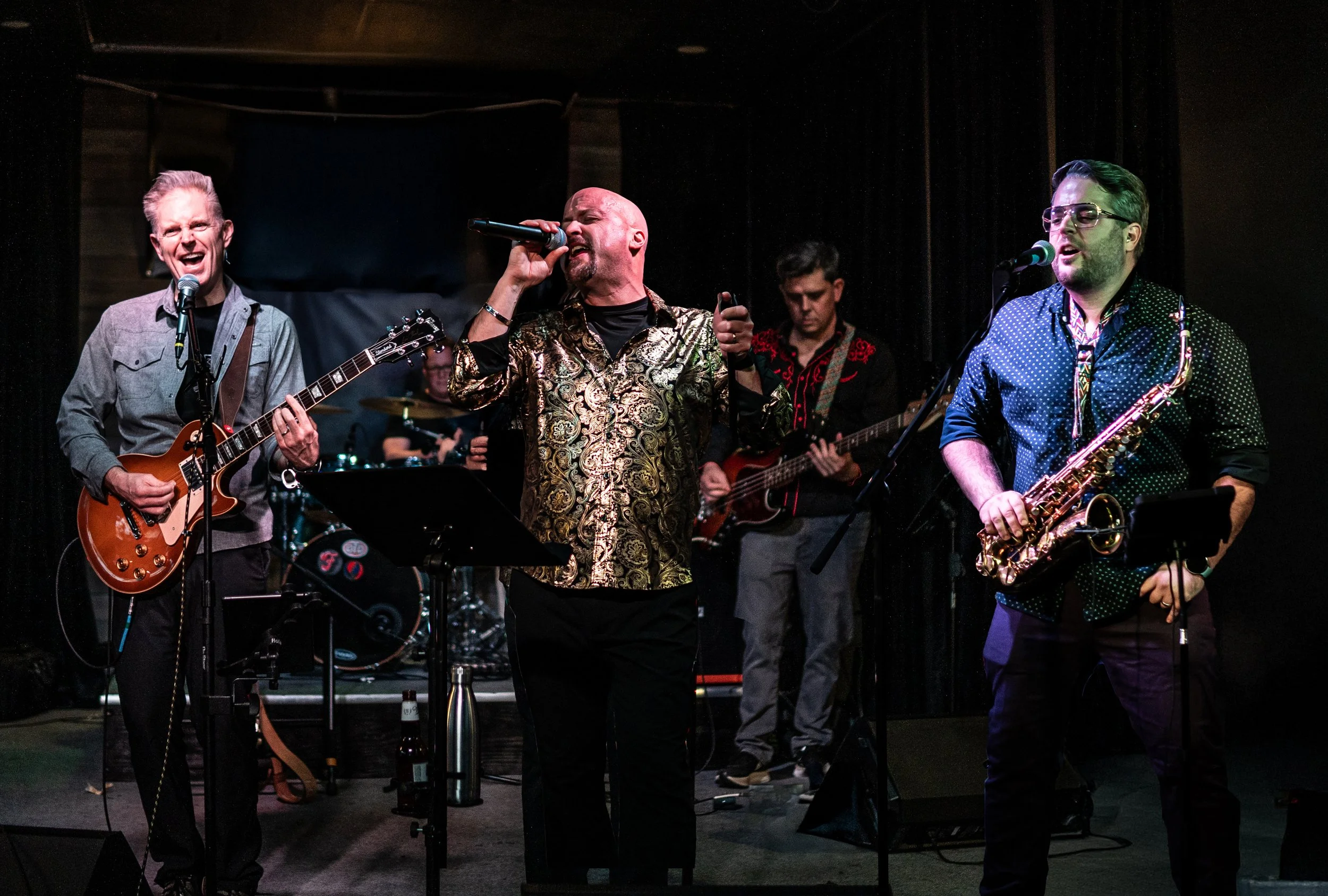 Band performing on stage with guitarist, vocalist, bassist, and saxophonist in a live music setting.