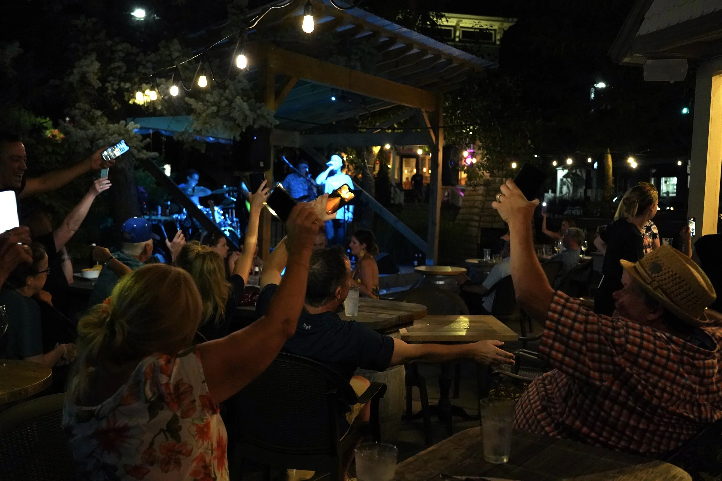 People gathered at an outdoor night music venue, some raising their glasses or phones, watching a live band perform on stage under string lights.