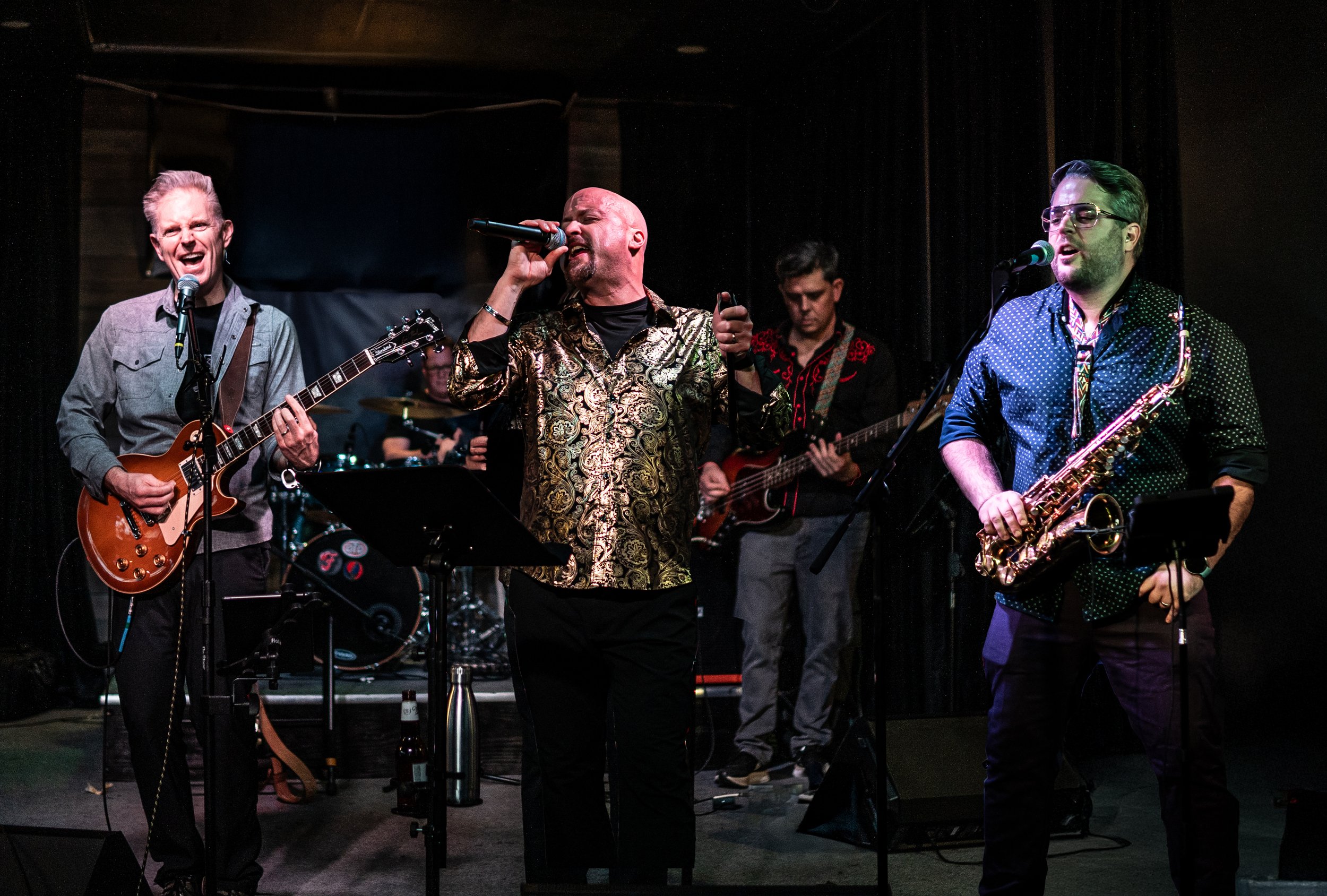 A band performing live on stage with four musicians: a guitarist on the left, a singer in the center, a bass player to the right, and a saxophonist on the far right, with a drummer partially visible in the background.