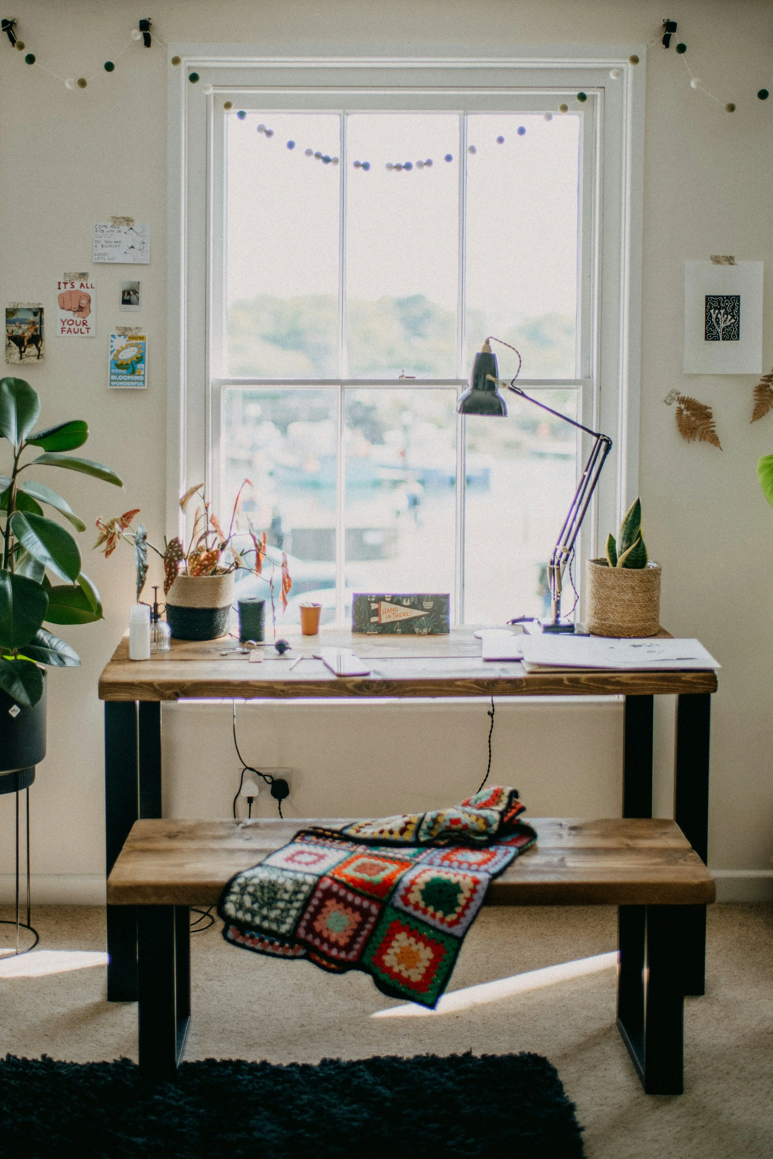 A modern workspace with a large wooden desk, potted plants on either side, a studio lamp, and a colorful crocheted blanket draped over the front edge. Behind the desk is a large window with a view of a harbor, decorated with string lights and small photos on the wall.