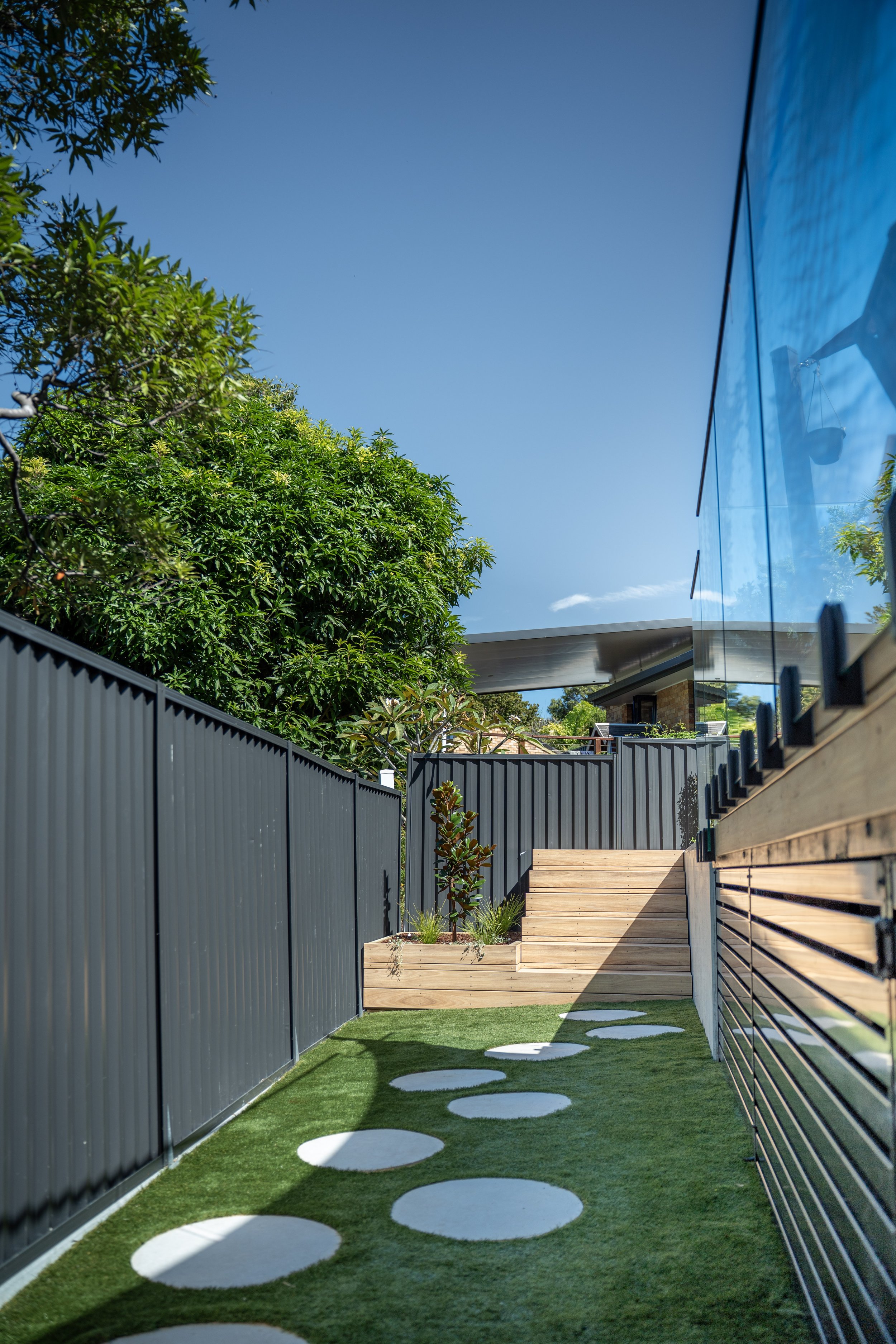 A modern backyard with a concrete pathway featuring circular stepping stones, bordered by a black metal fence, with a grassy area and a wooden deck with stairs on the right side. A glass building with reflection is visible on the right, and lush green trees are on the left under a clear blue sky.