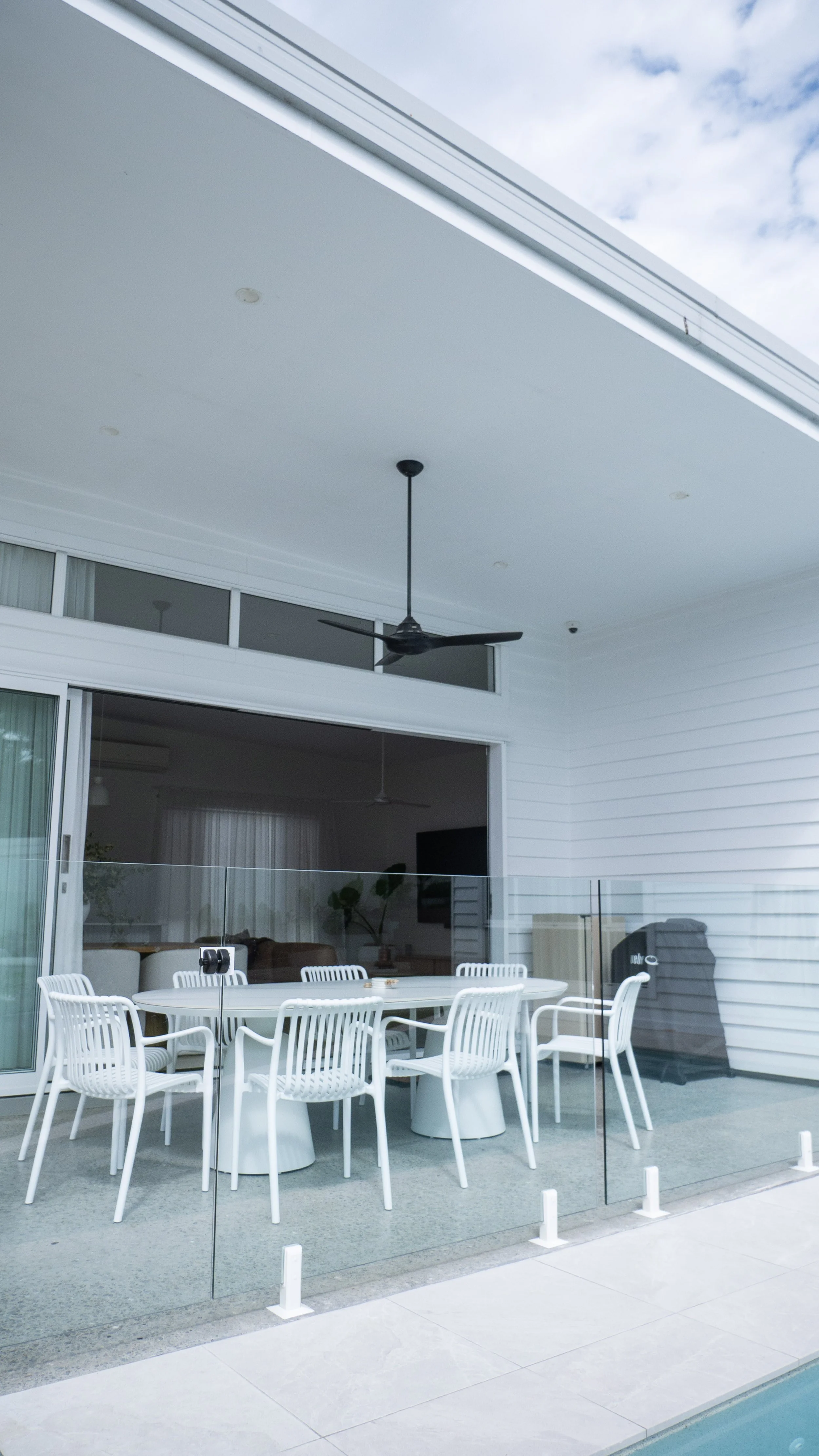 Outdoor covered patio with white table and chairs, glass railing, ceiling fans, sliding glass door, and partial view of interior living room, exterior wall with white siding, and cloudy sky.