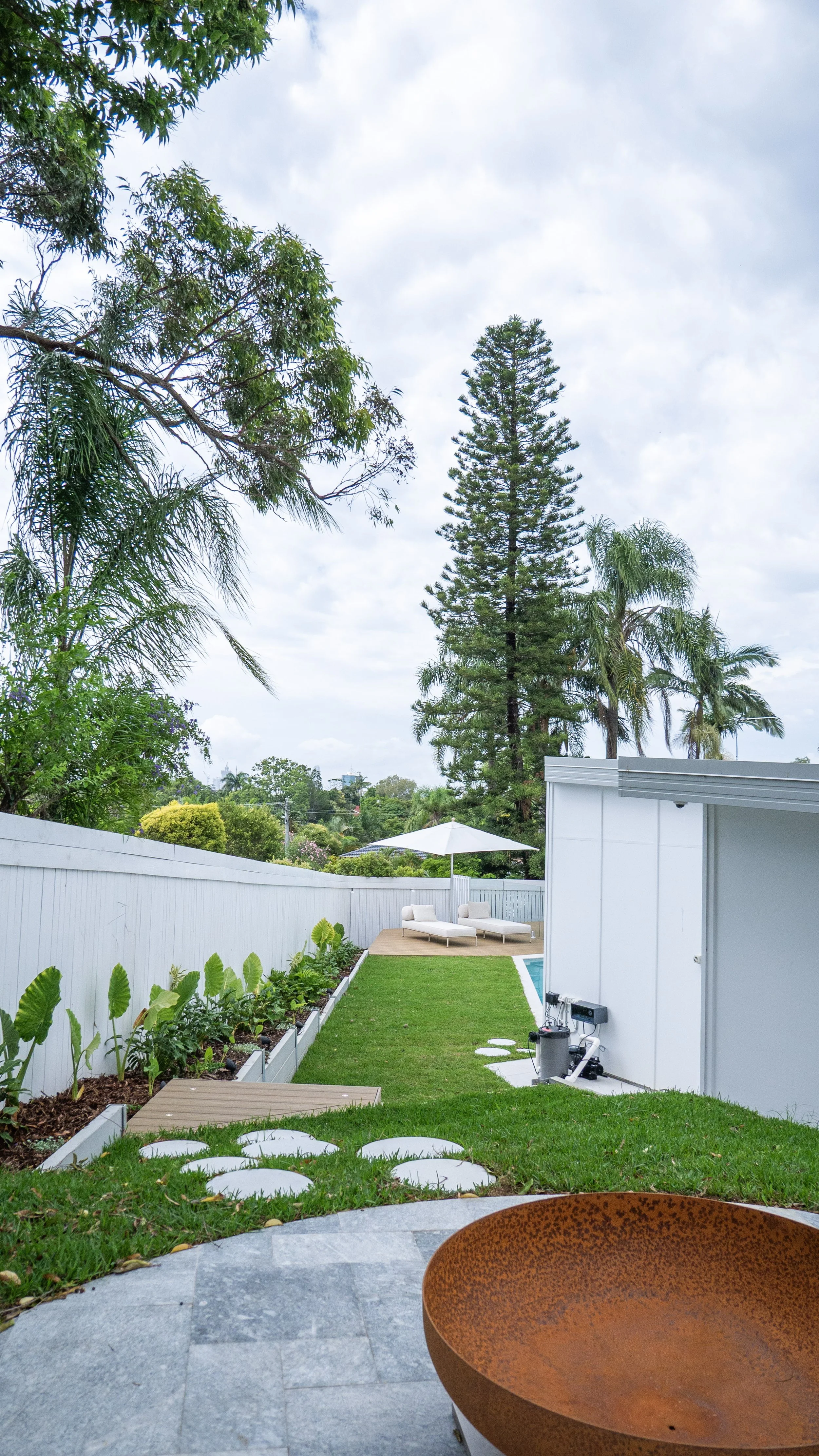 A backyard with a swimming pool, a white outdoor table with two lounge chairs, green plants, a white fence, a large tall pine tree, and partly cloudy sky.