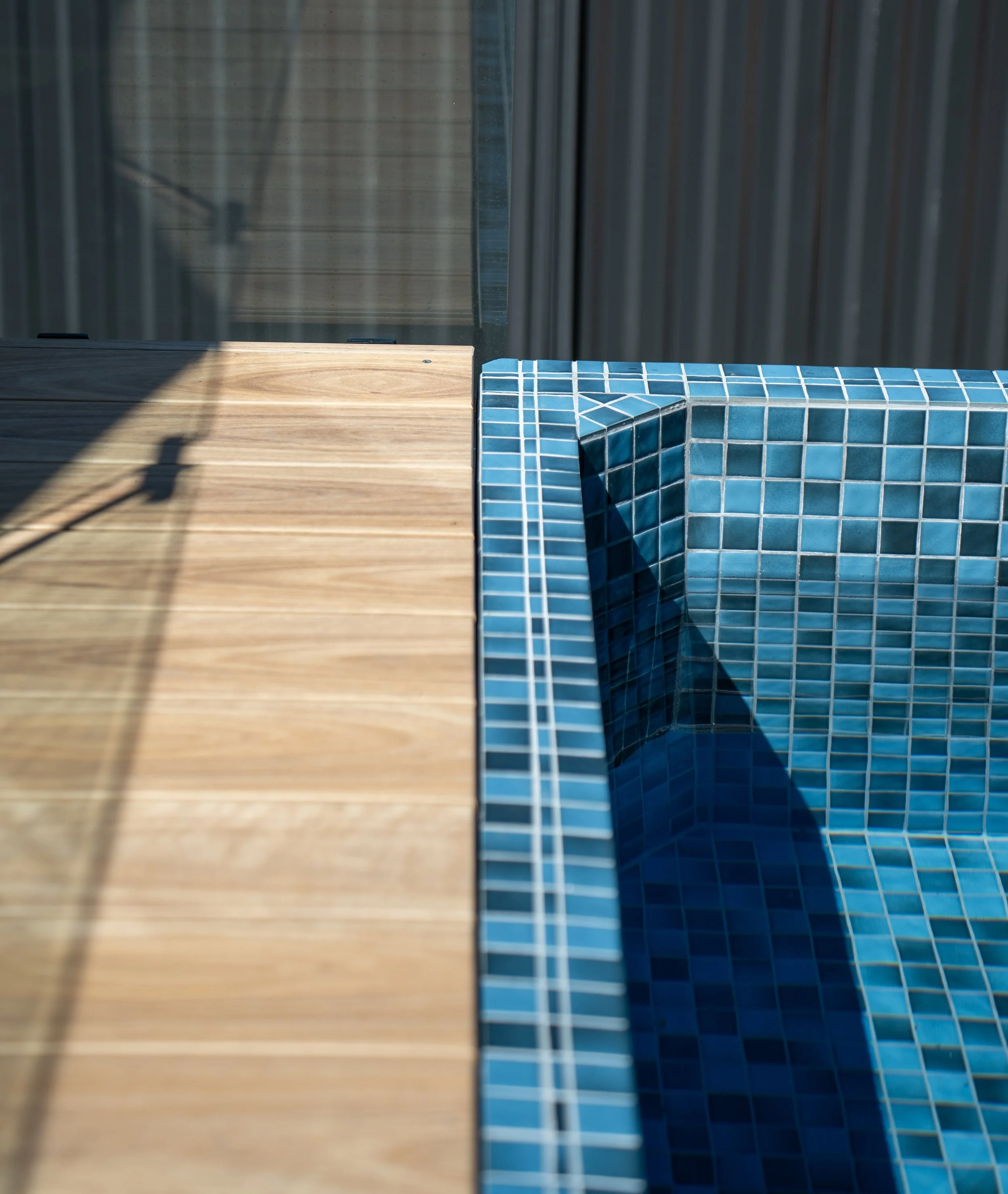 Close-up of a swimming pool with a tiled edge and wooden deck, shadow cast on the deck.