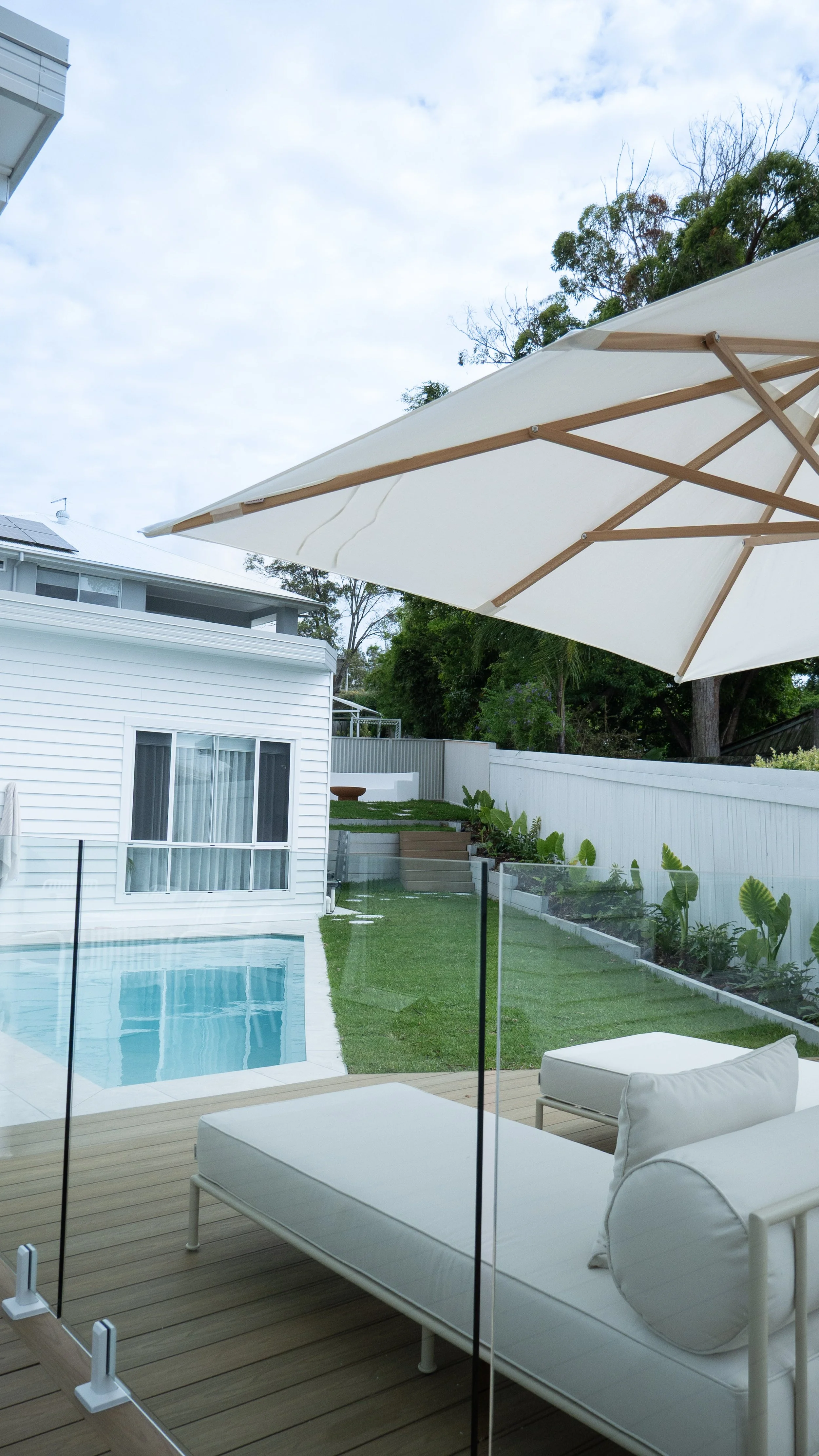 Outdoor pool area with lounge chair, large white patio umbrella, and backyard yard with garden and trees, surrounded by a white fence, and a blue sky with clouds.