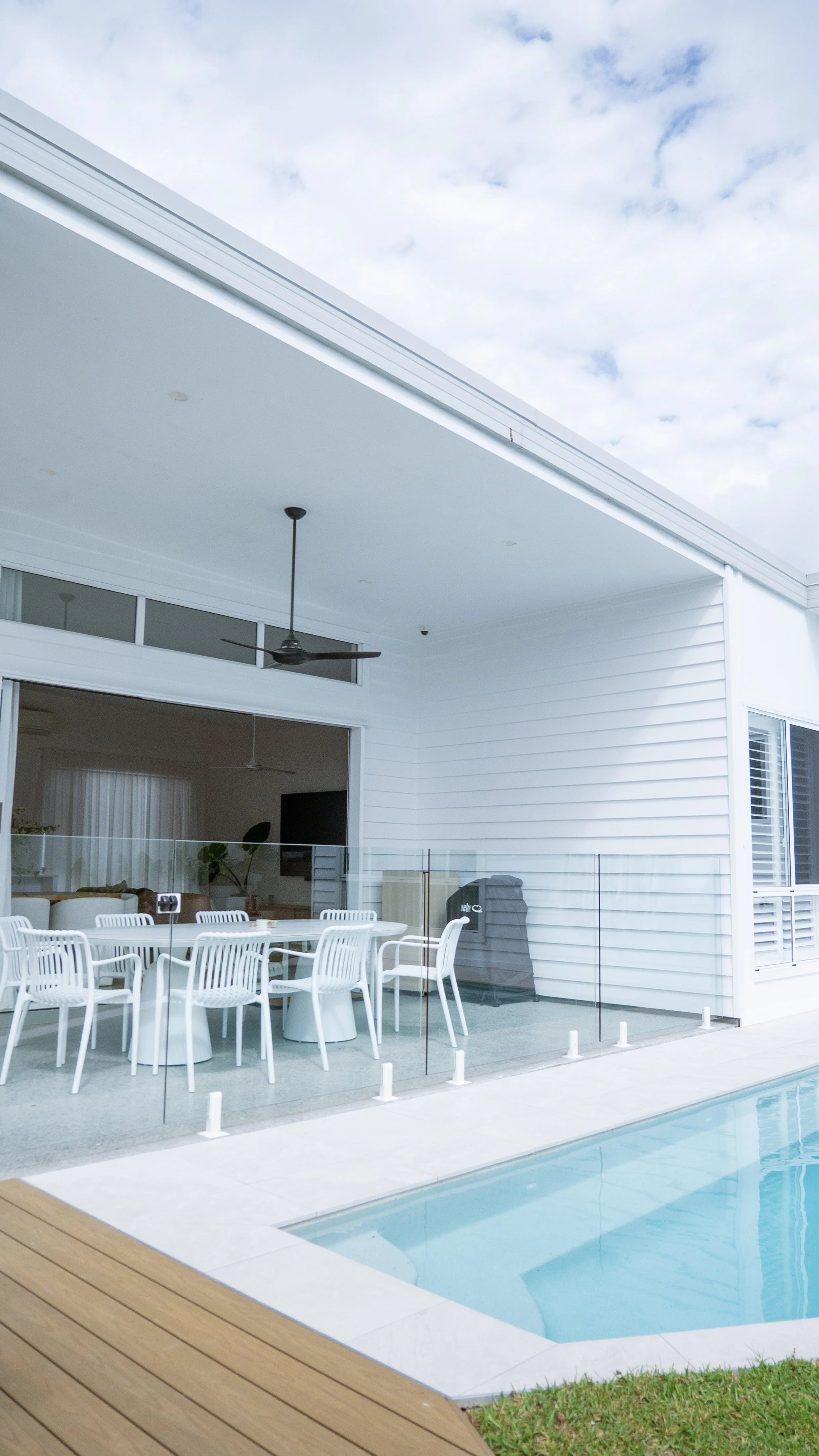 Modern outdoor patio with white dining table and chairs, glass barrier, swimming pool, and contemporary house with white siding and large windows, under a partly cloudy sky.