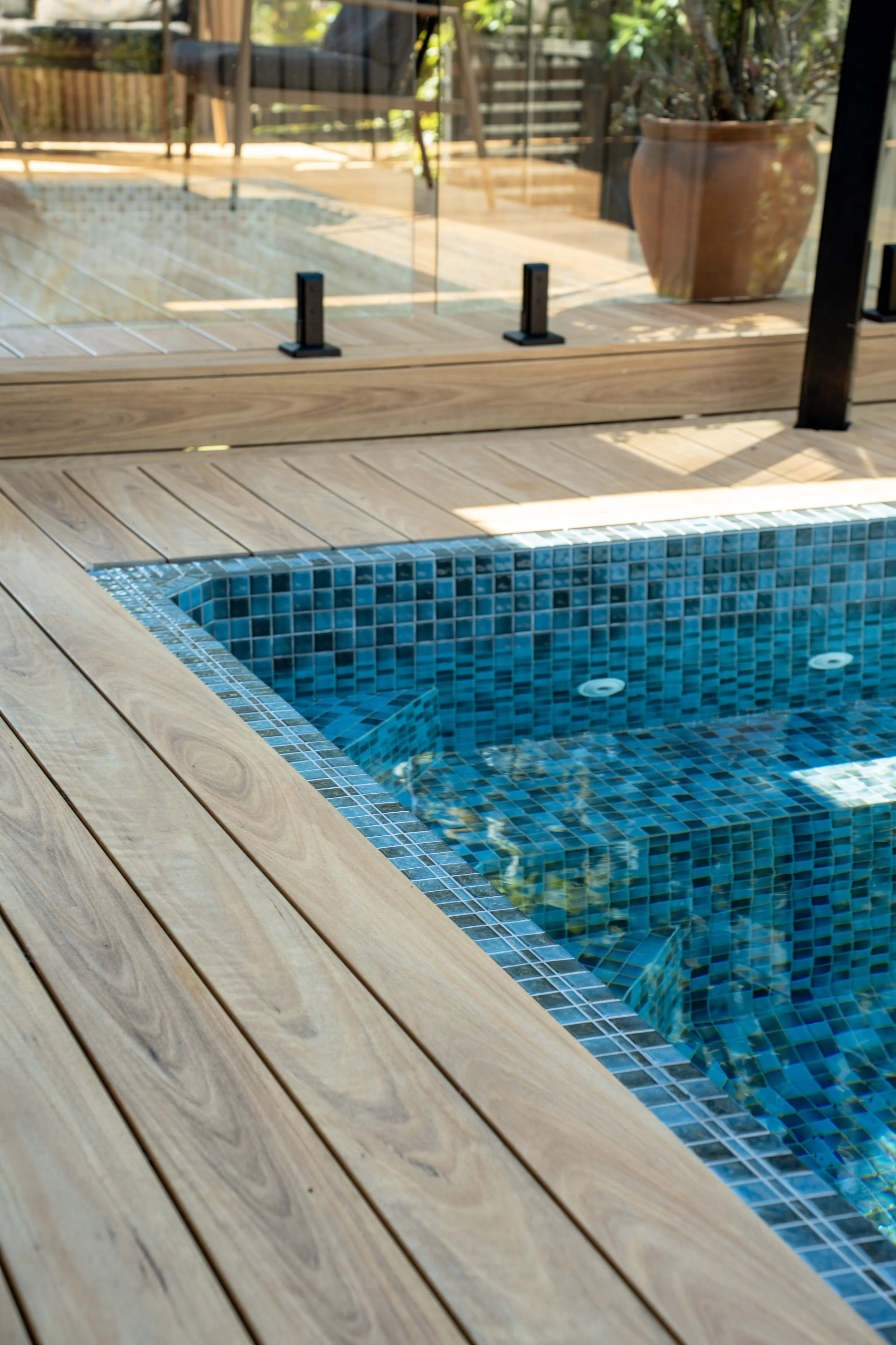 A backyard scene showing a swimming pool with blue mosaic tiles, surrounded by a wooden deck and a glass safety fence, with potted plants in the background.