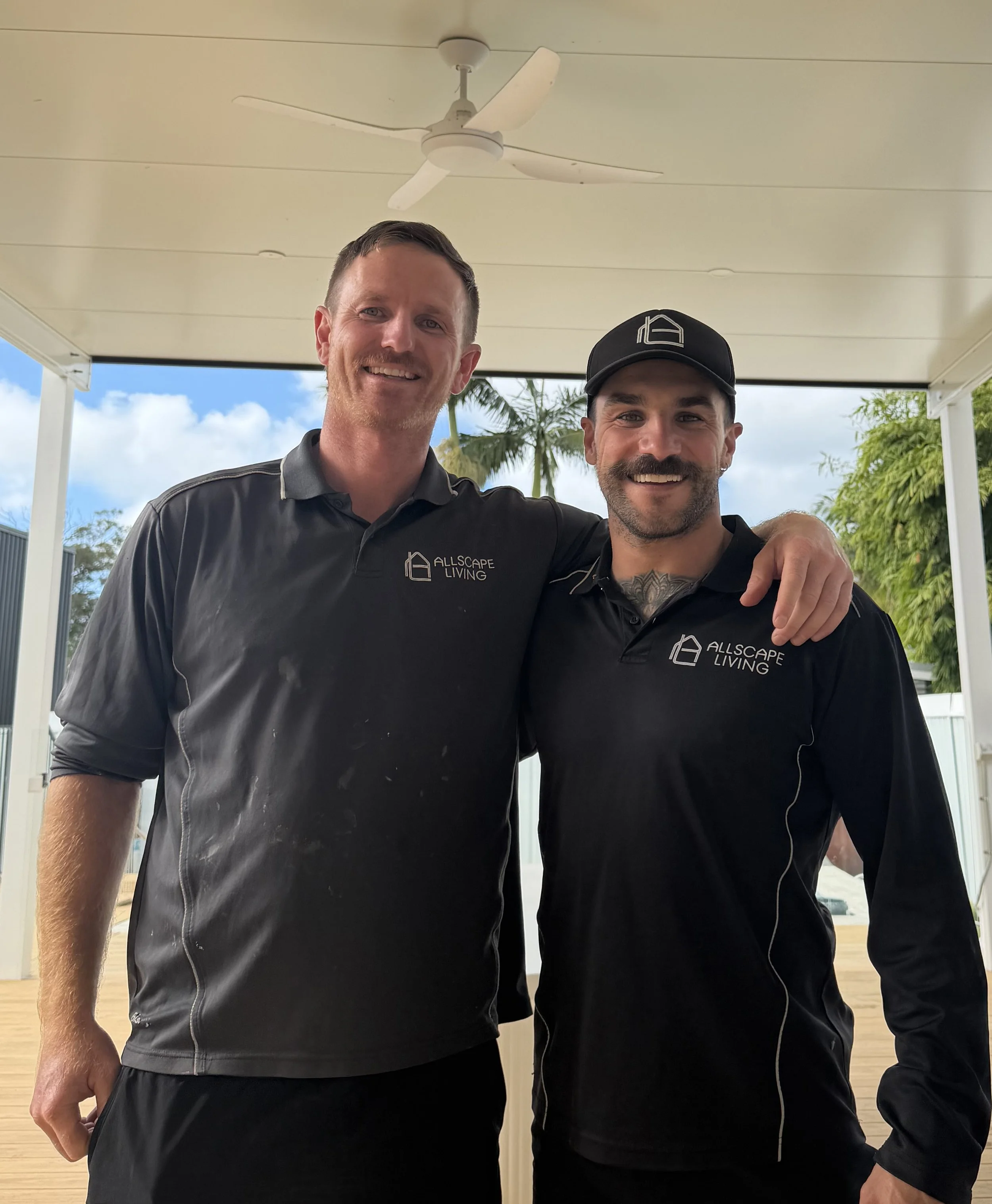 Two smiling men with facial hair and short hair standing close together under a ceiling fan. Both wear black shirts with a logo that says 'Allscape Living'. They are outside with trees and blue sky visible in the background.