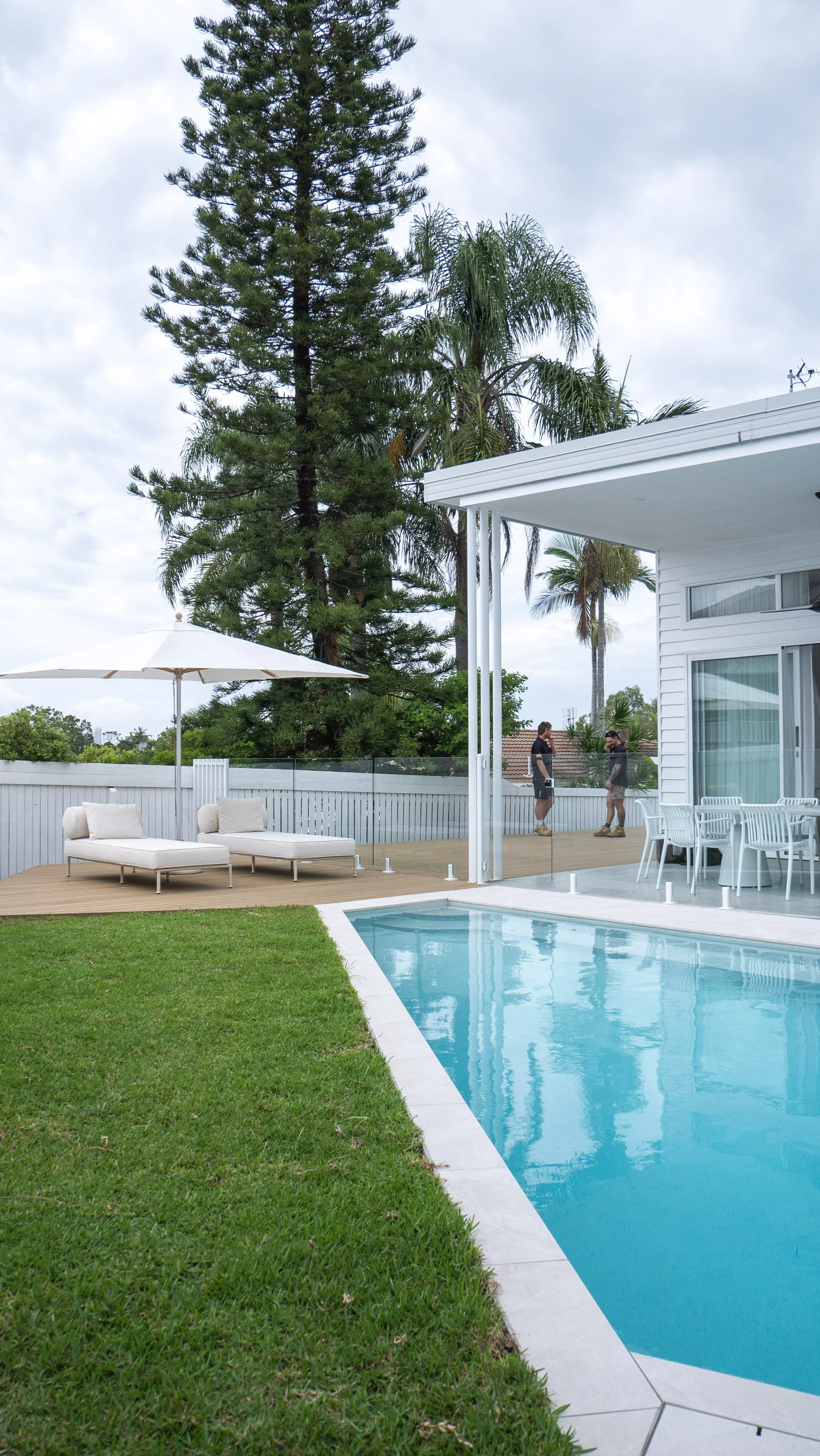 View of a backyard with a swimming pool, green grass, outdoor furniture, a white fence, tall trees, and a patio with people talking.