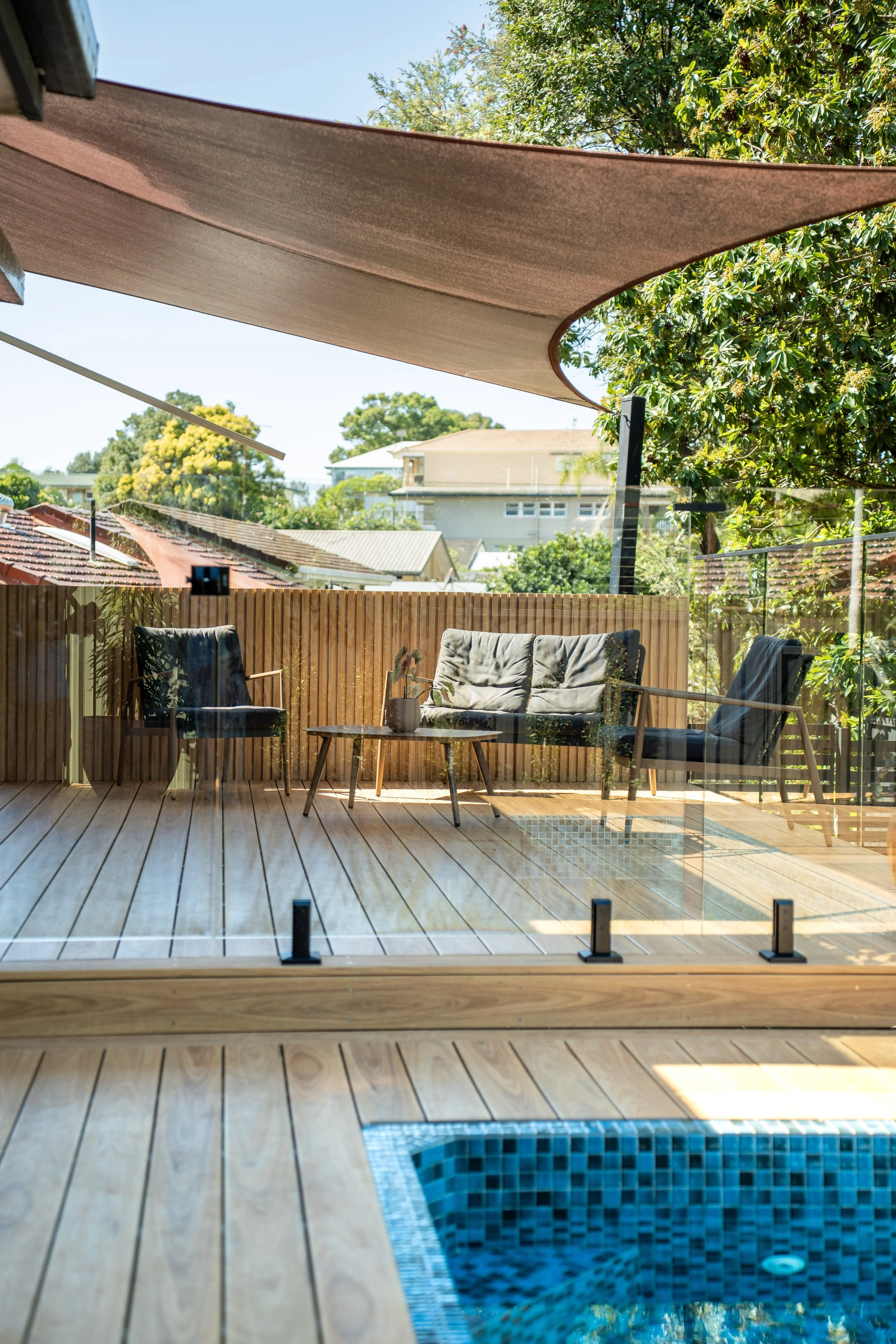 Backyard patio with outdoor seating and shade sail, overlooking neighboring residential buildings and trees, with a small pool in the foreground.