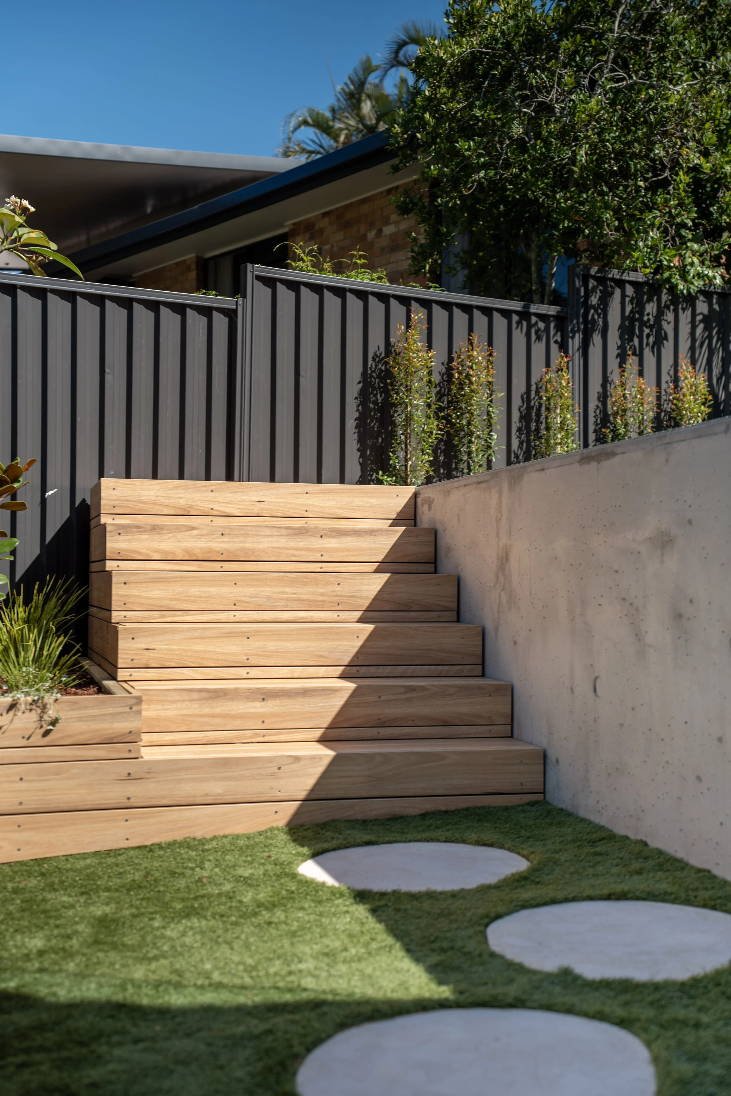 Outdoor scene with wooden stairs leading up to a modern house, green grass area with circular stepping stones, a gray metal fence, and a large leafy tree beside the house.