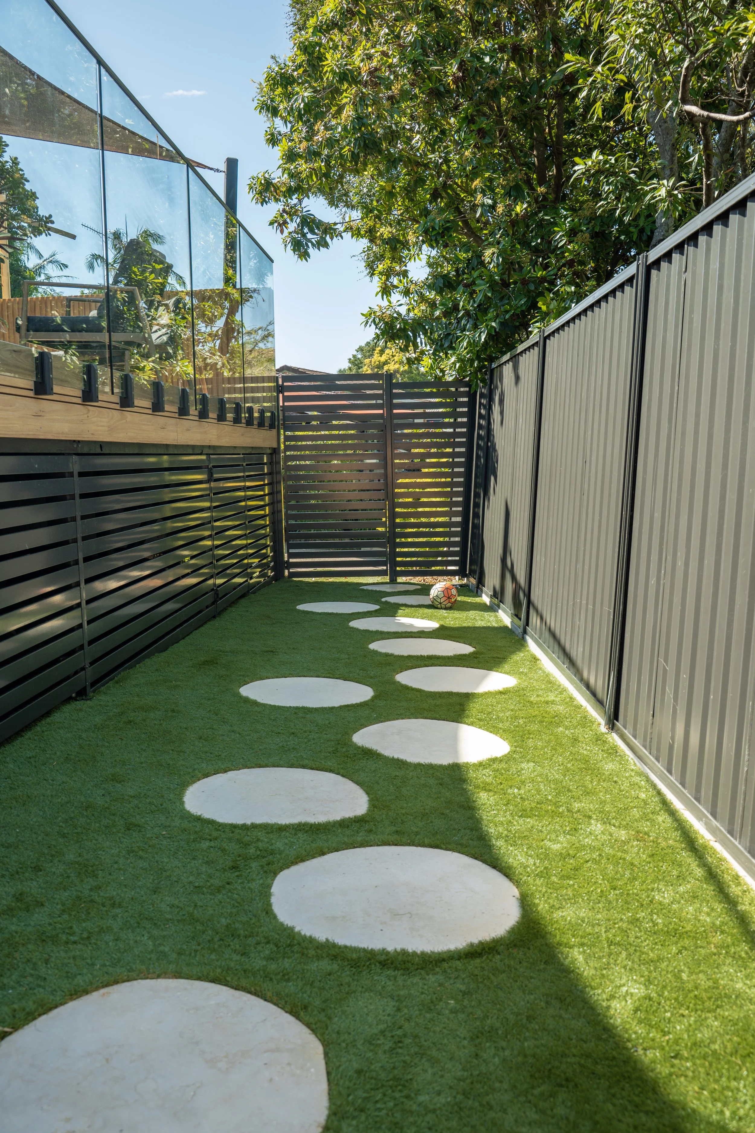 A narrow backyard with green artificial grass and white stepping stones leading to a black gate, surrounded by black metal fence, with a large tree and neighboring house with a deck on the left.