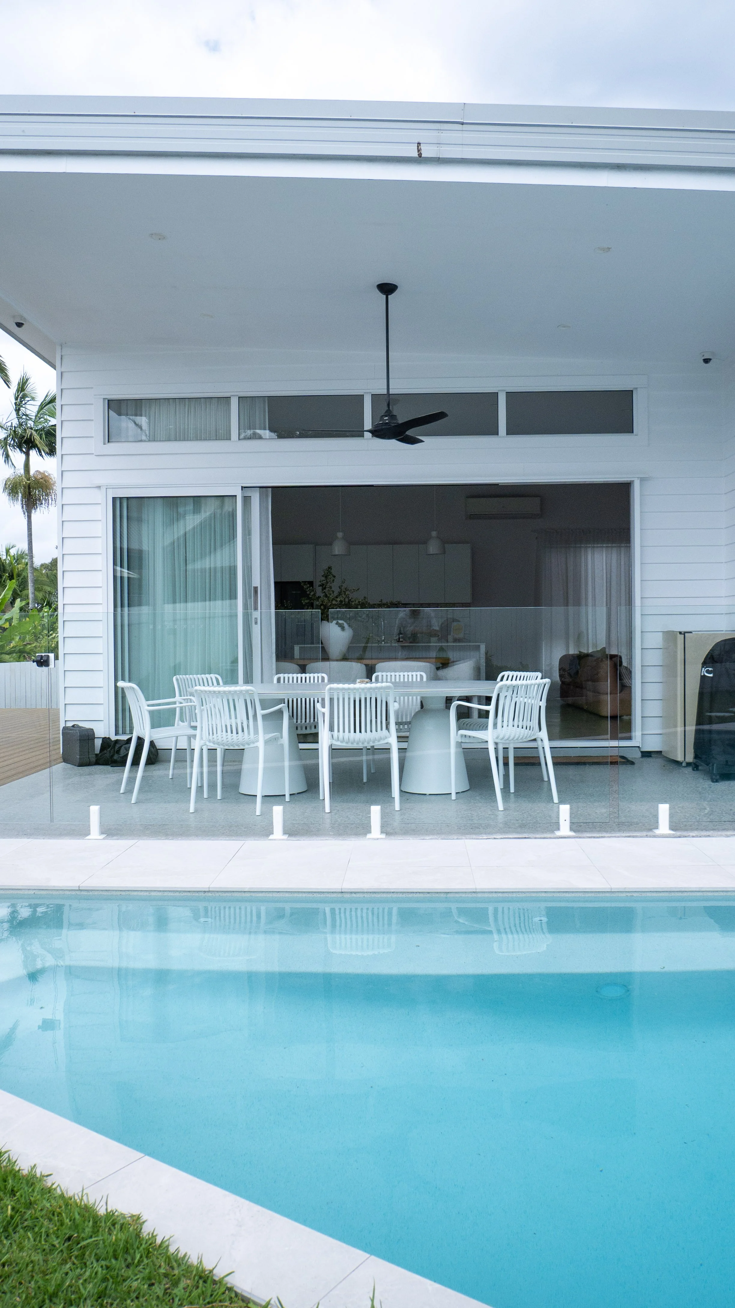 Modern house with sliding glass doors opening to a patio with white outdoor furniture. A pool is in the foreground, and the interior of the house features a kitchen and living area visible through the glass.
