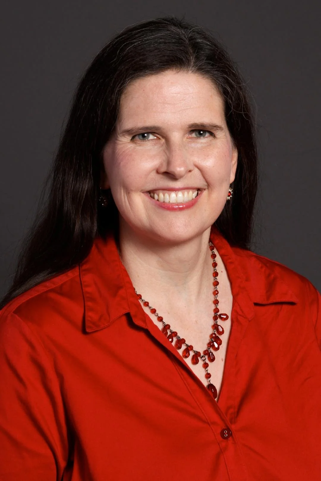 A woman with long dark hair, wearing a red blouse and matching red jewelry, smiling in a professional studio portrait with a dark background.