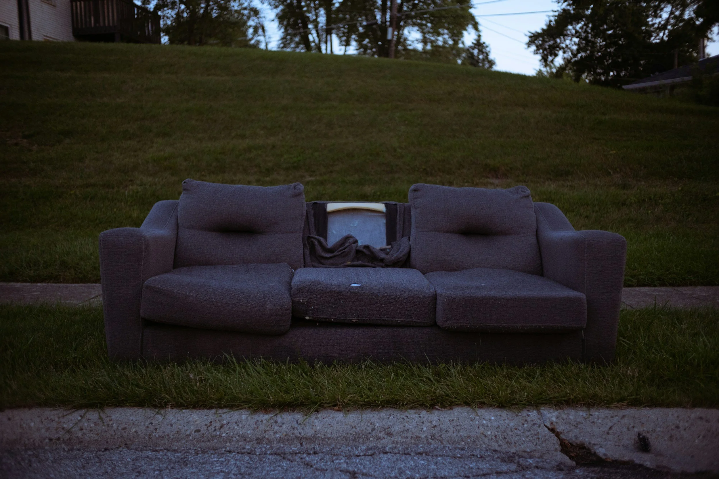 A dark outdoor scene featuring a worn gray couch with pillows, placed on grass in front of a grassy hill with trees and a house in the background.