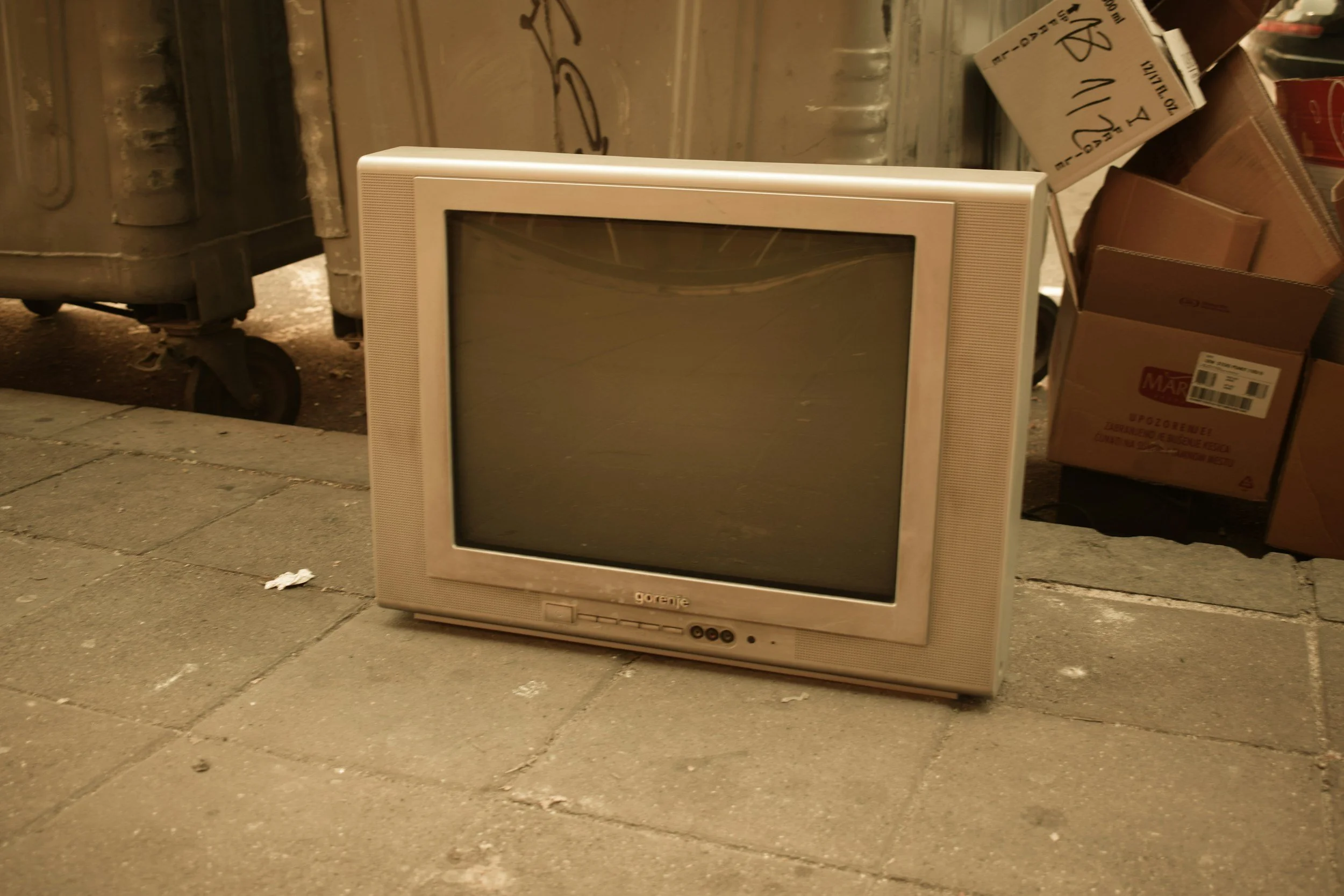 An old CRT television with a grayish-white frame on a sidewalk, next to cardboard boxes and trash cans.