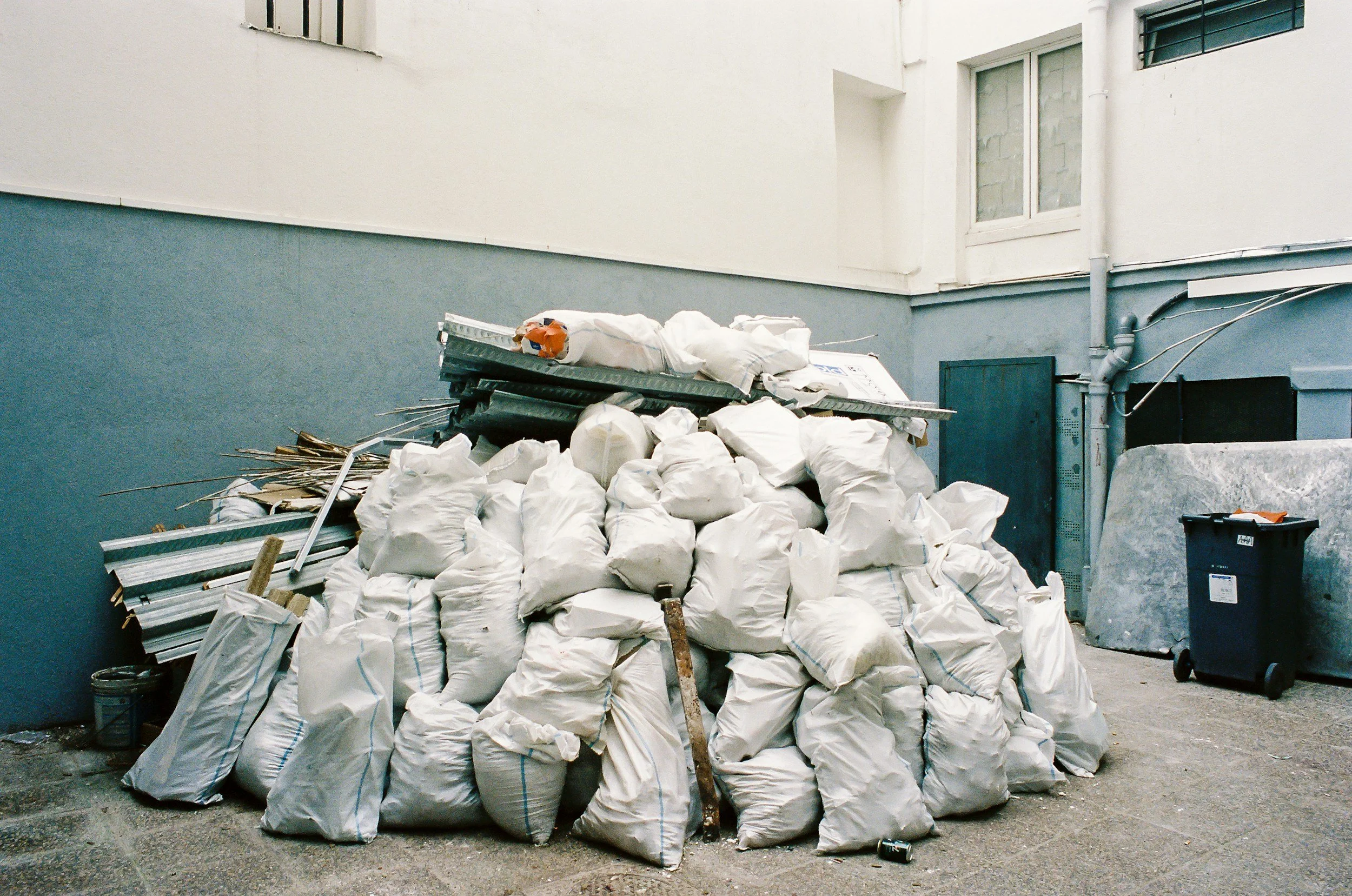 A pile of white trash bags and debris, including wooden sticks and metal pieces, sitting outside near a building with a two-tone wall and scattered pipes.