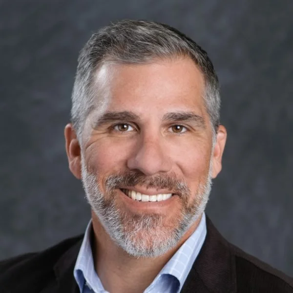 A smiling man with short gray hair and a beard, wearing a blue shirt and a dark blazer, posing for a professional headshot against a dark background.
