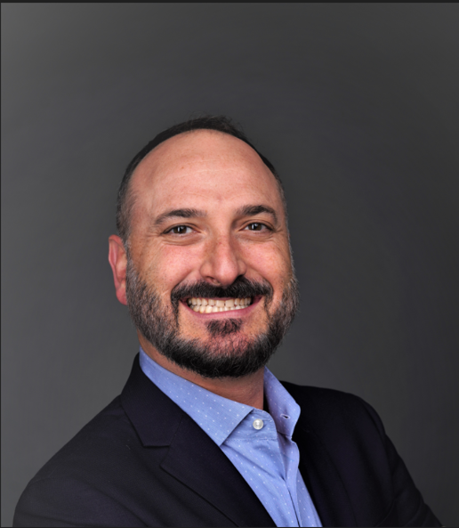 A man with a beard and short hair smiling, wearing a navy blazer and light blue dress shirt against a gray background.
