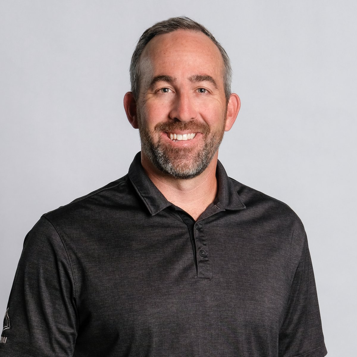 Portrait of a smiling man with short gray hair and a beard, wearing a black polo shirt, against a plain light gray background.