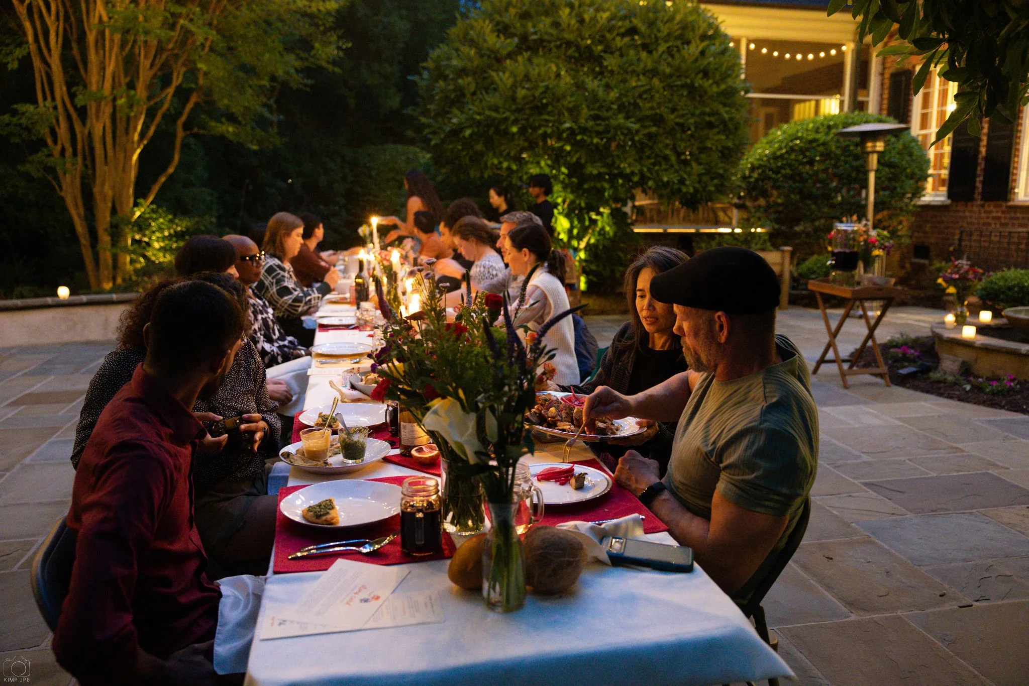 A group of people gathered around a long dinner table outdoors during evening. They are enjoying a meal with plates of food, surrounded by decorative flowers and candles, with a house and string lights in the background.