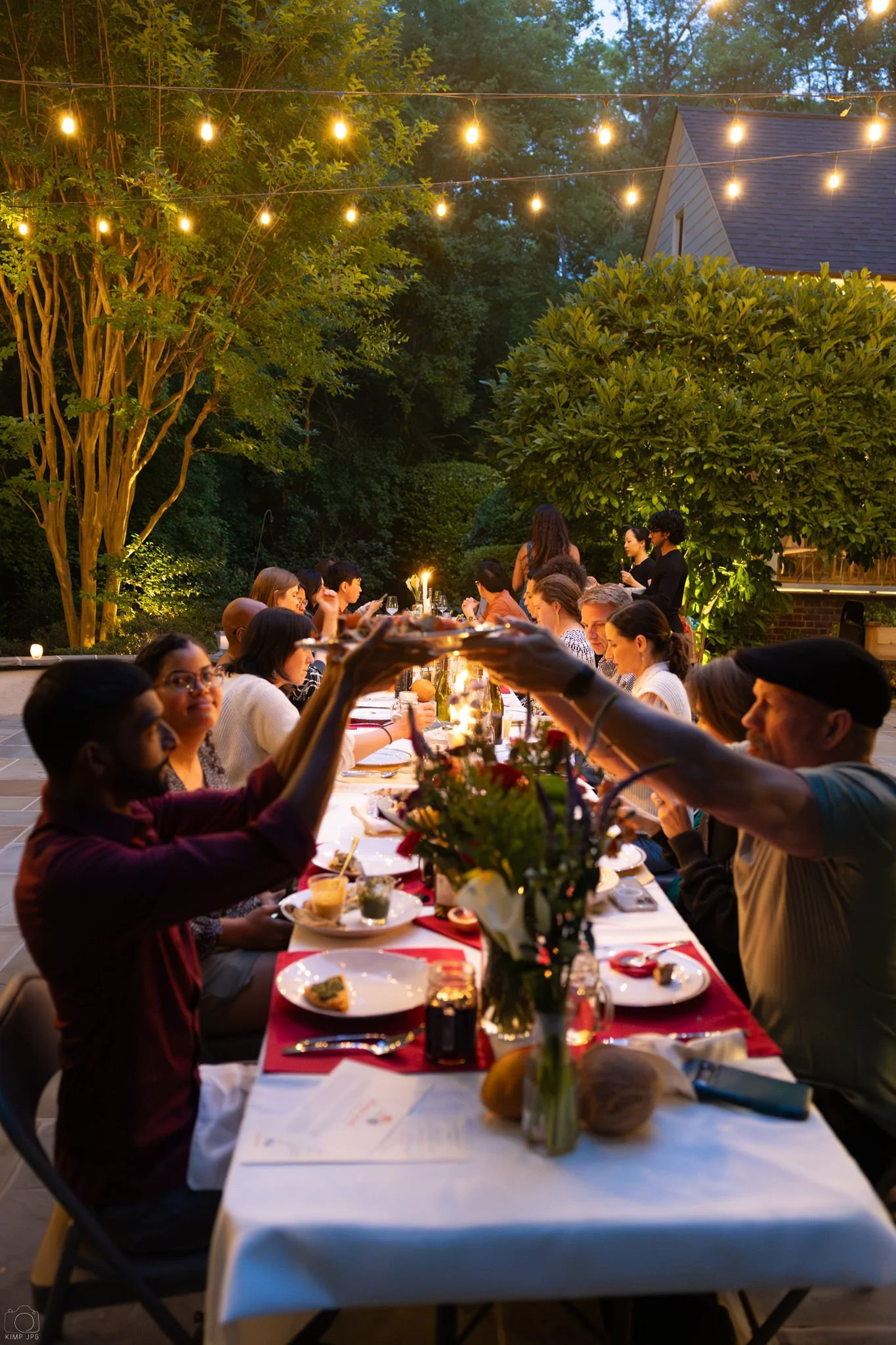 People gathered around a long outdoor dinner table during evening, decorated with flowers, candles, and surrounded by string lights in a backyard garden.