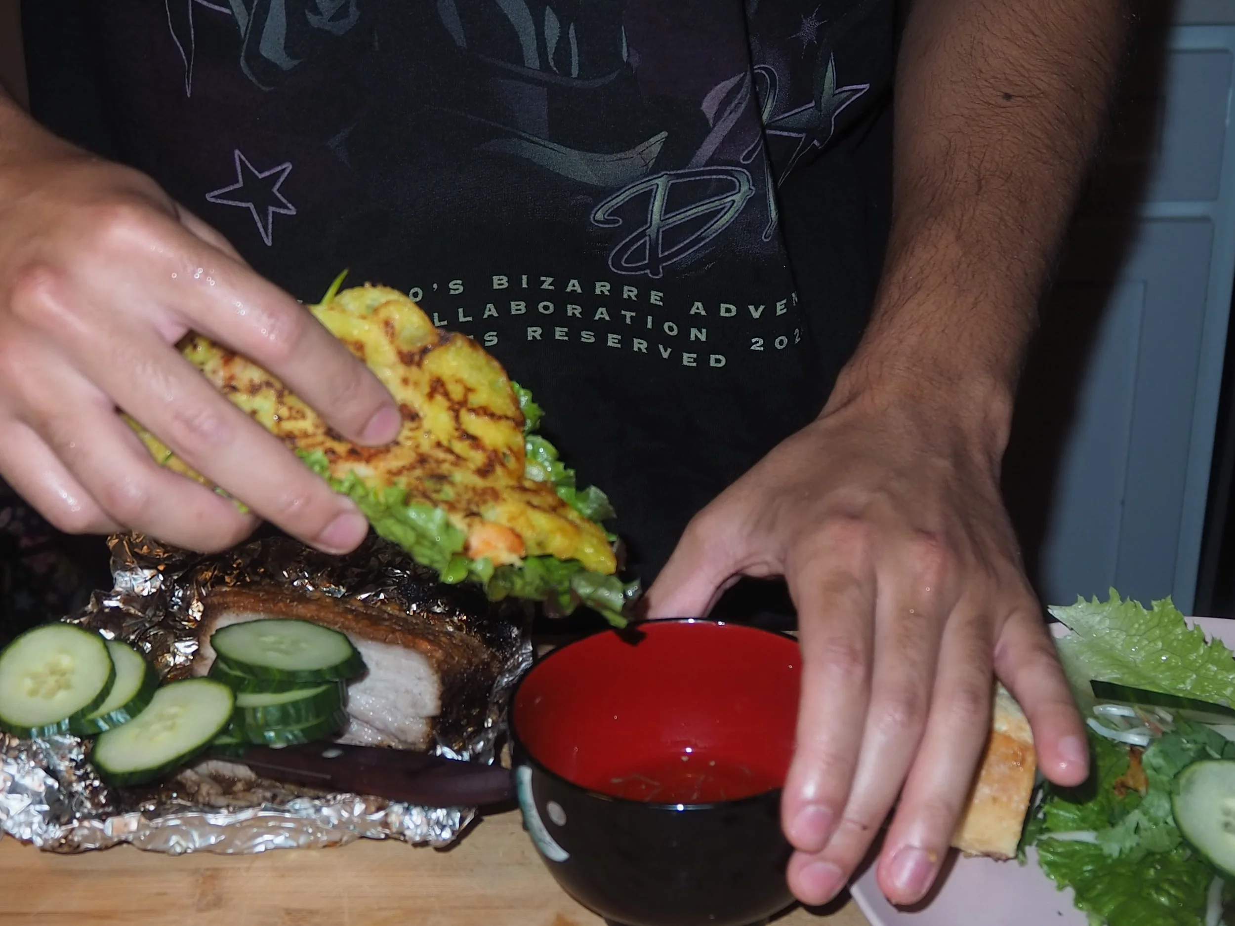Person assembling a sandwich with sliced cucumbers, lettuce, grilled chicken, and grilled pineapple on a cutting board with foil-wrapped foil and a red container.