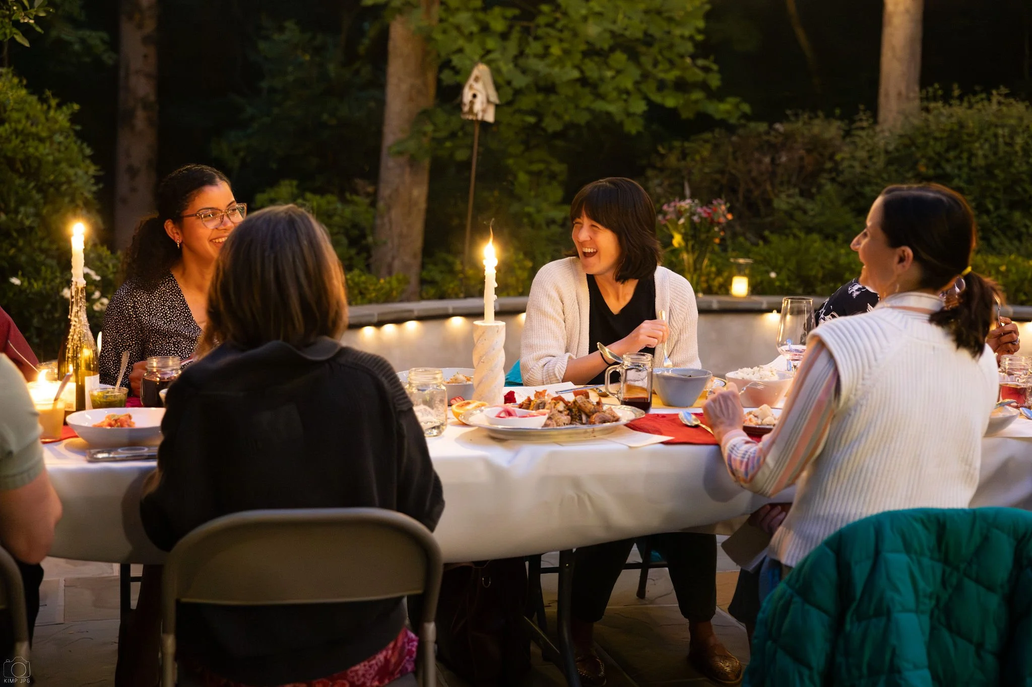 People sitting around a table outdoors at night, enjoying a meal and laughter, with trees and hanging decorative birdhouse in the background.
