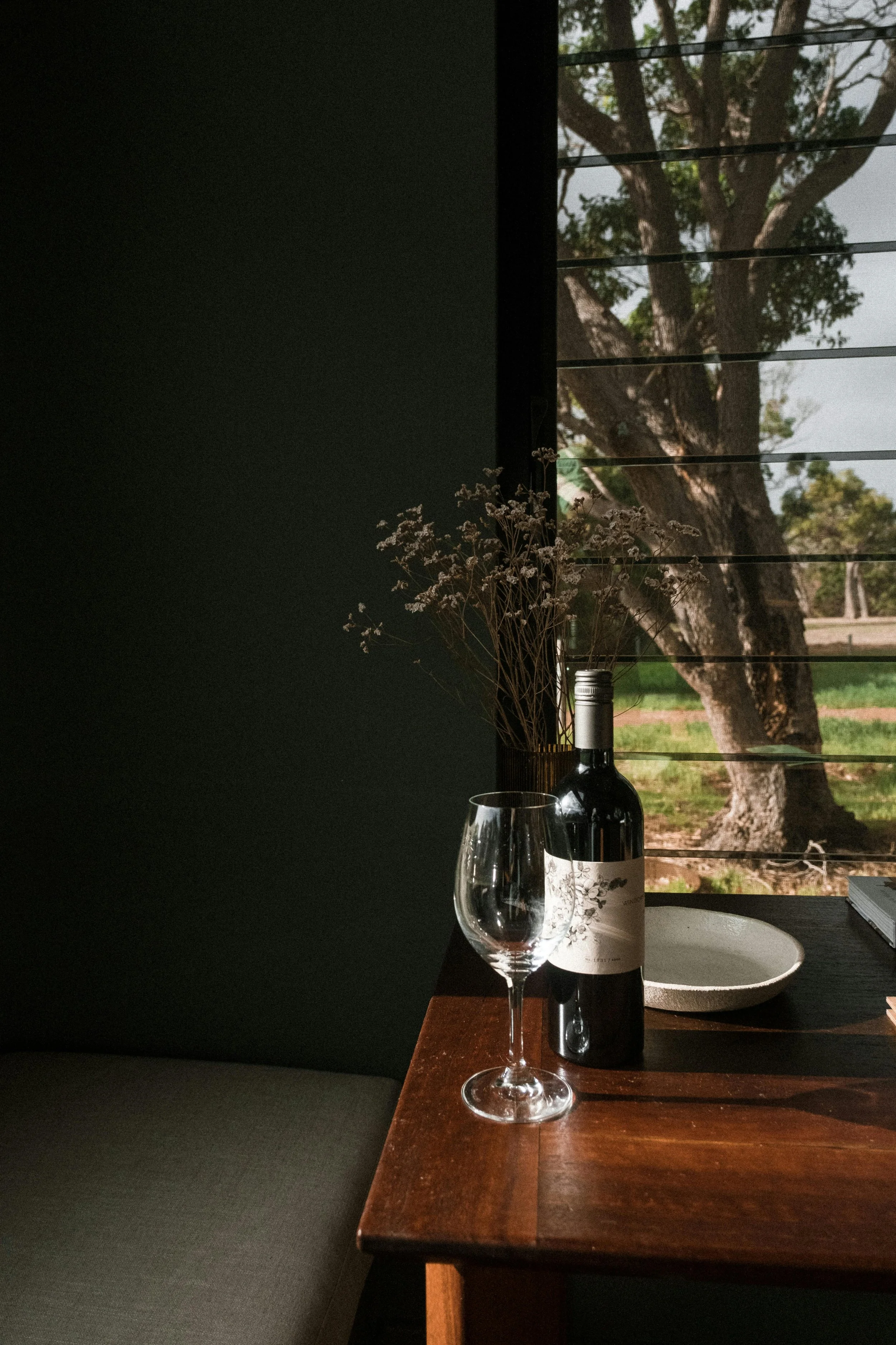 A wooden table with a wine bottle, empty wine glass, a white ceramic bowl, and a book, with a window showing trees outside in the background.