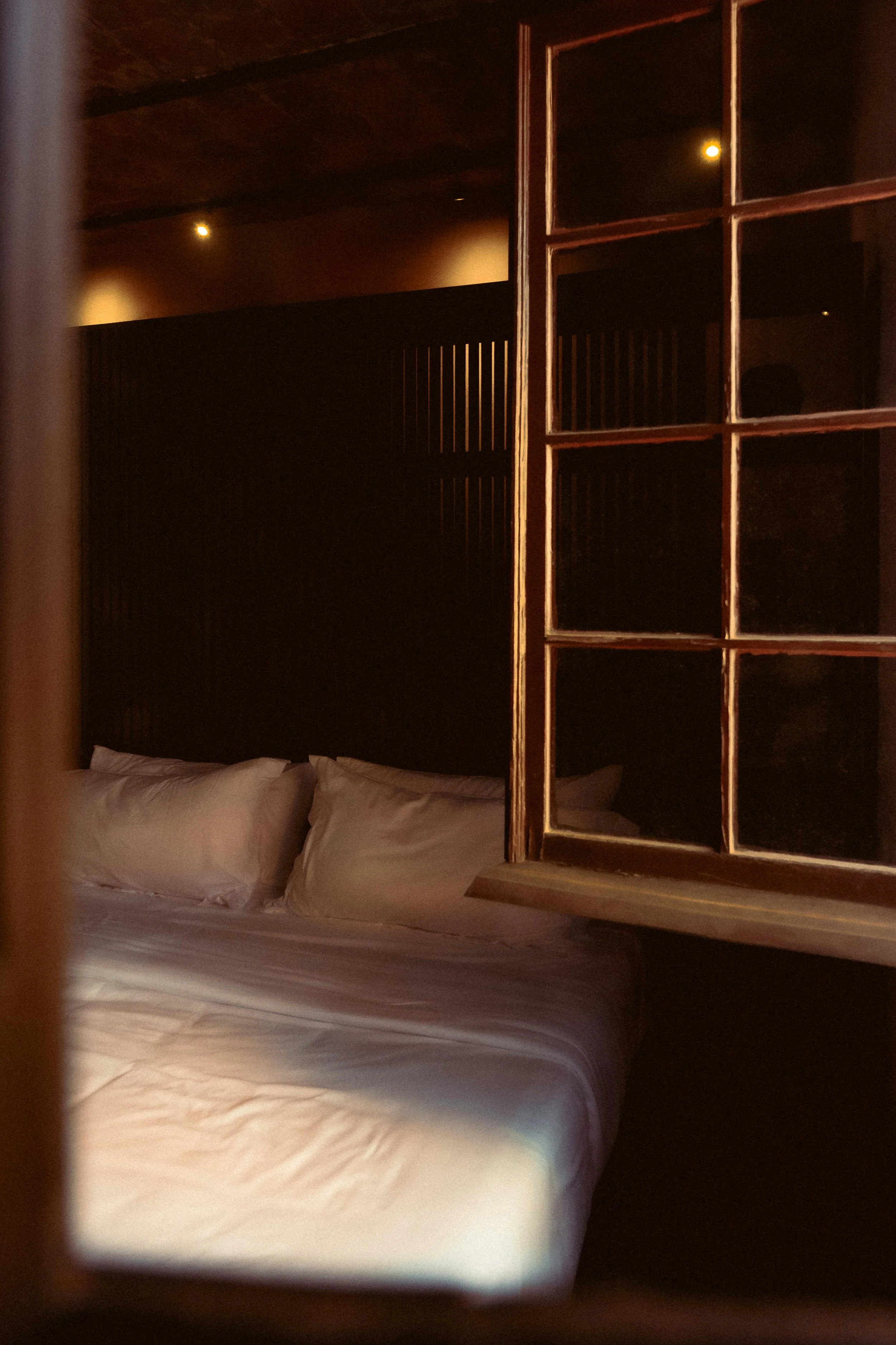 A view of a dark bedroom seen through a wooden window with glass panes. There are pillows on a bed with white linens and soft lighting.