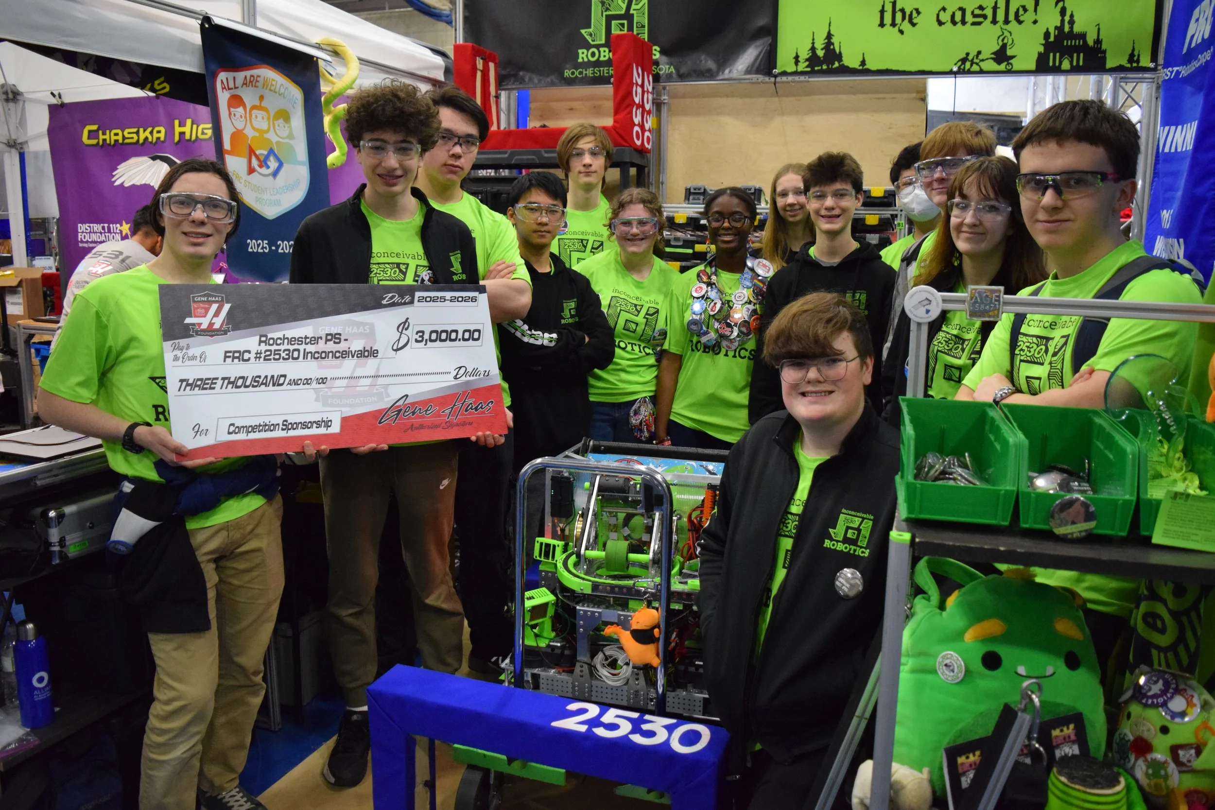 A group of young students at a robotics competition, wearing matching bright green shirts and safety glasses, standing behind their robot and a large check for $3,000. The background features banners and equipment related to robotics.