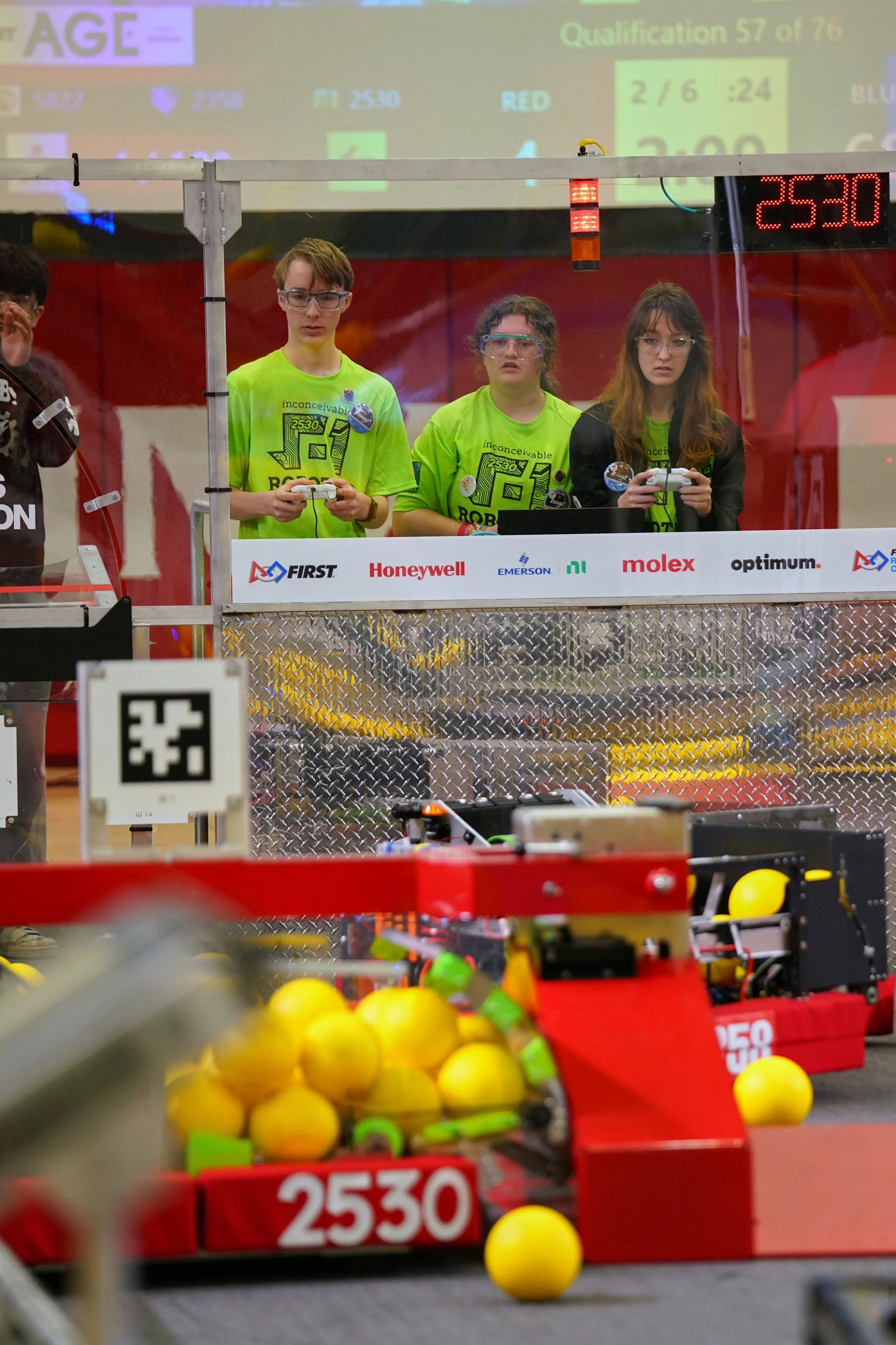 Three young people wearing bright green shirts and safety glasses are operating a robotics competition robot behind a glass partition. The background shows a digital scoreboard and a projection screen with scores and qualification information. The foreground has a bin filled with yellow balls, and a red container with the number 2530.