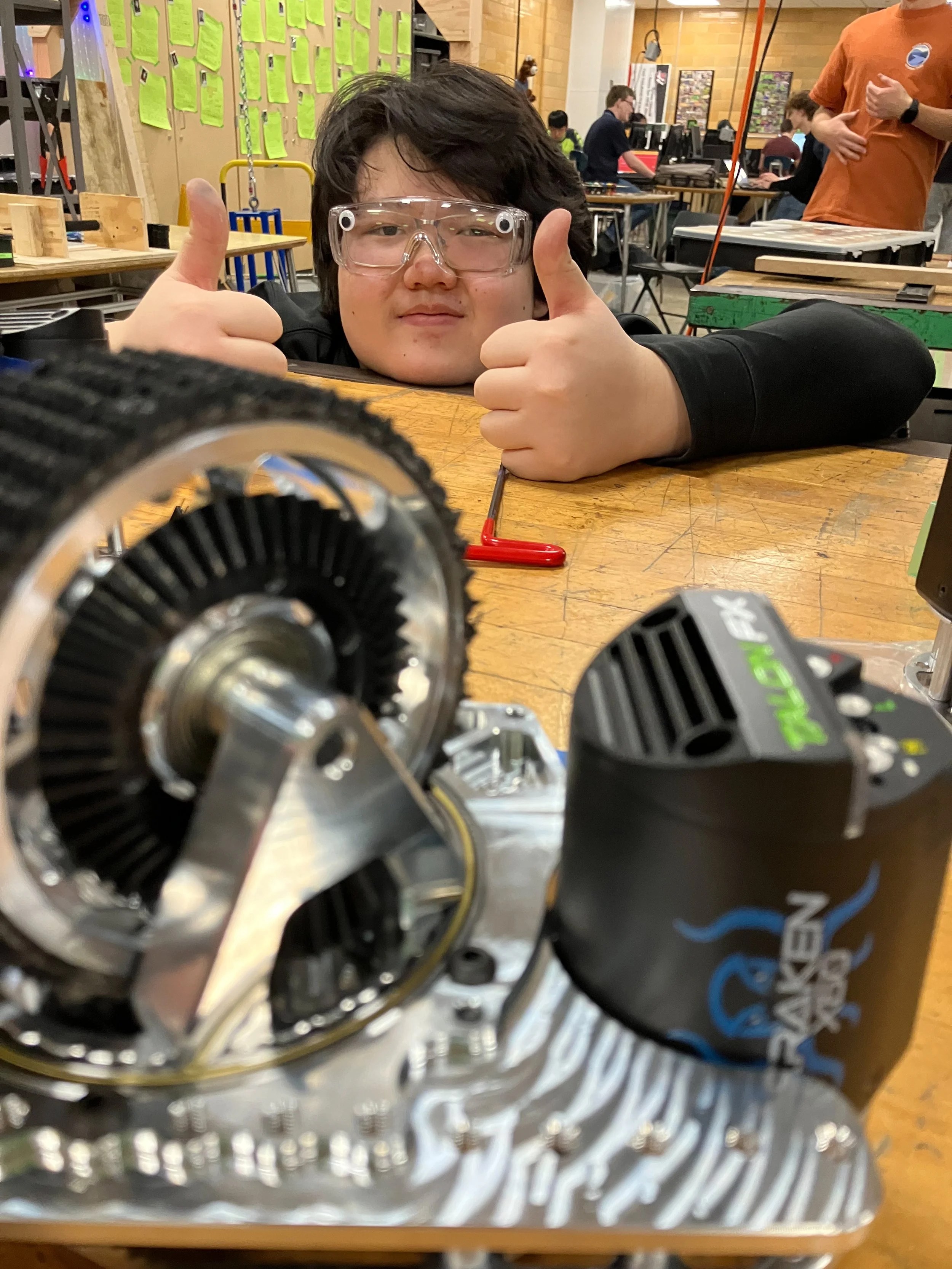 Person wearing safety glasses resting their head on a workbench, giving a thumbs-up in a workshop with various mechanical parts and tools around.