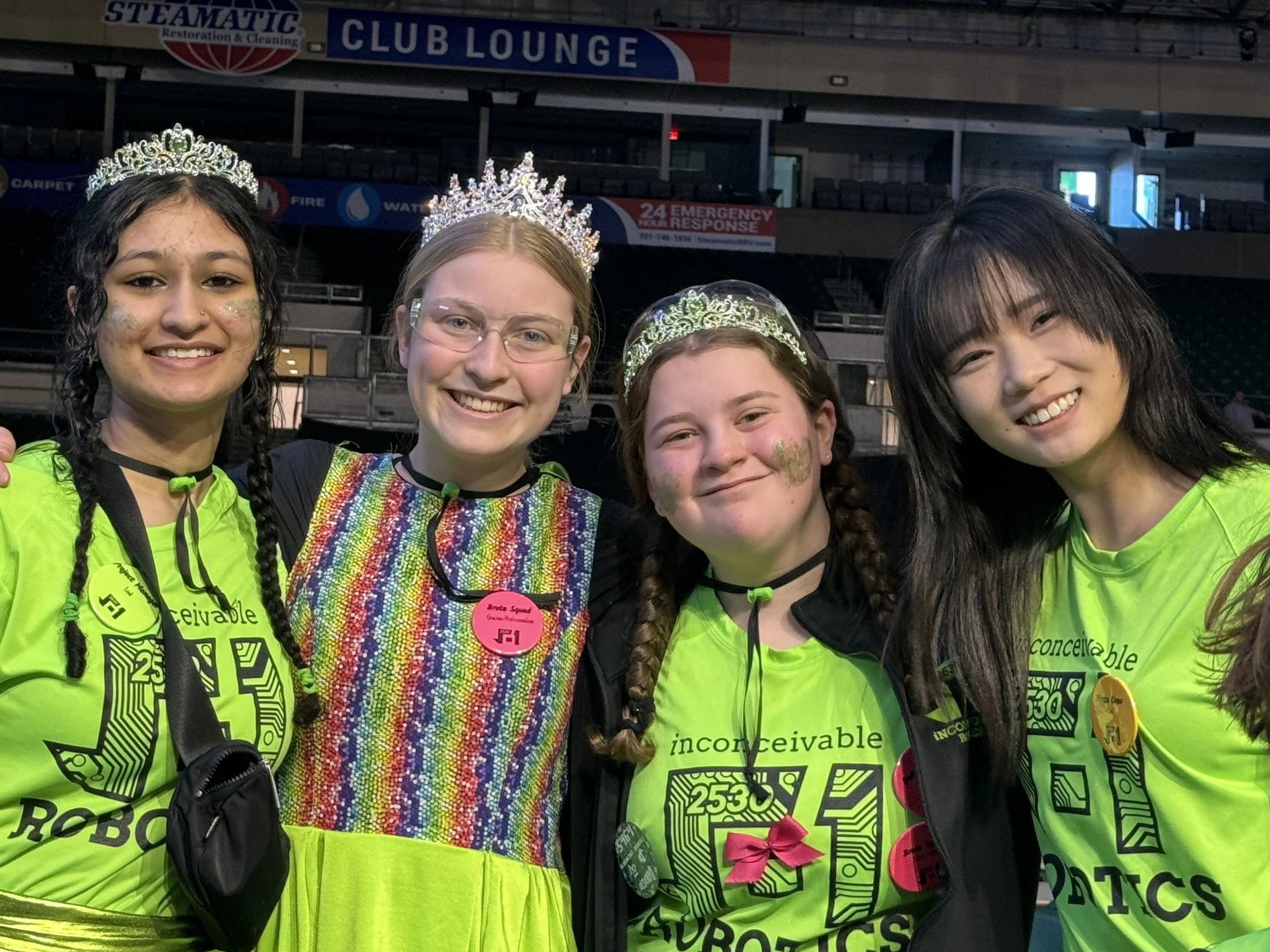 Four young women wearing tiaras and bright yellow-green shirts, standing together and smiling at the camera, in an indoor arena with signs and banners in the background.