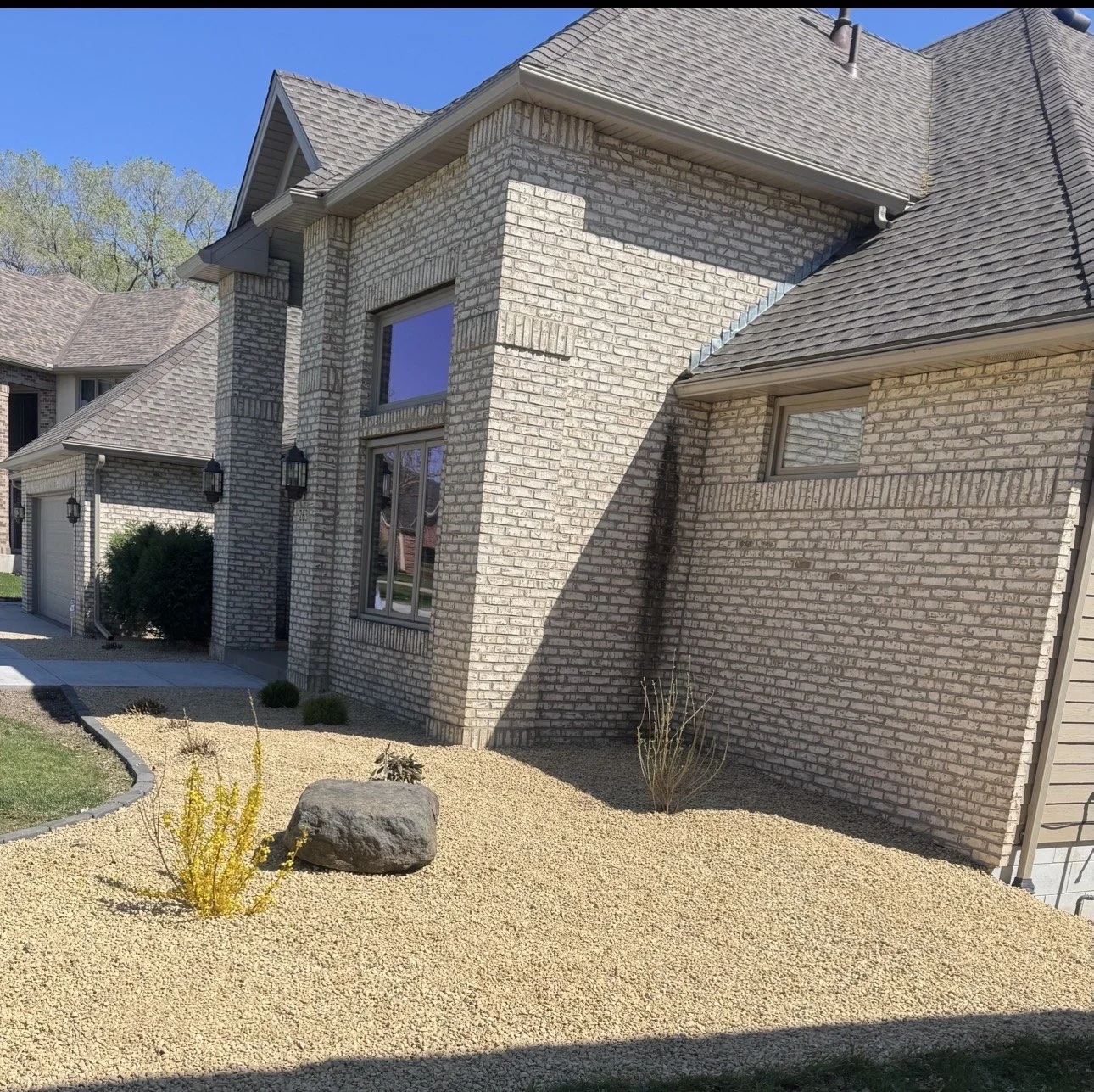 Front view of a modern house with a brick exterior and sloped roof, featuring a small landscaped yard with gravel, a large rock, and some sparse plants, under a clear blue sky.