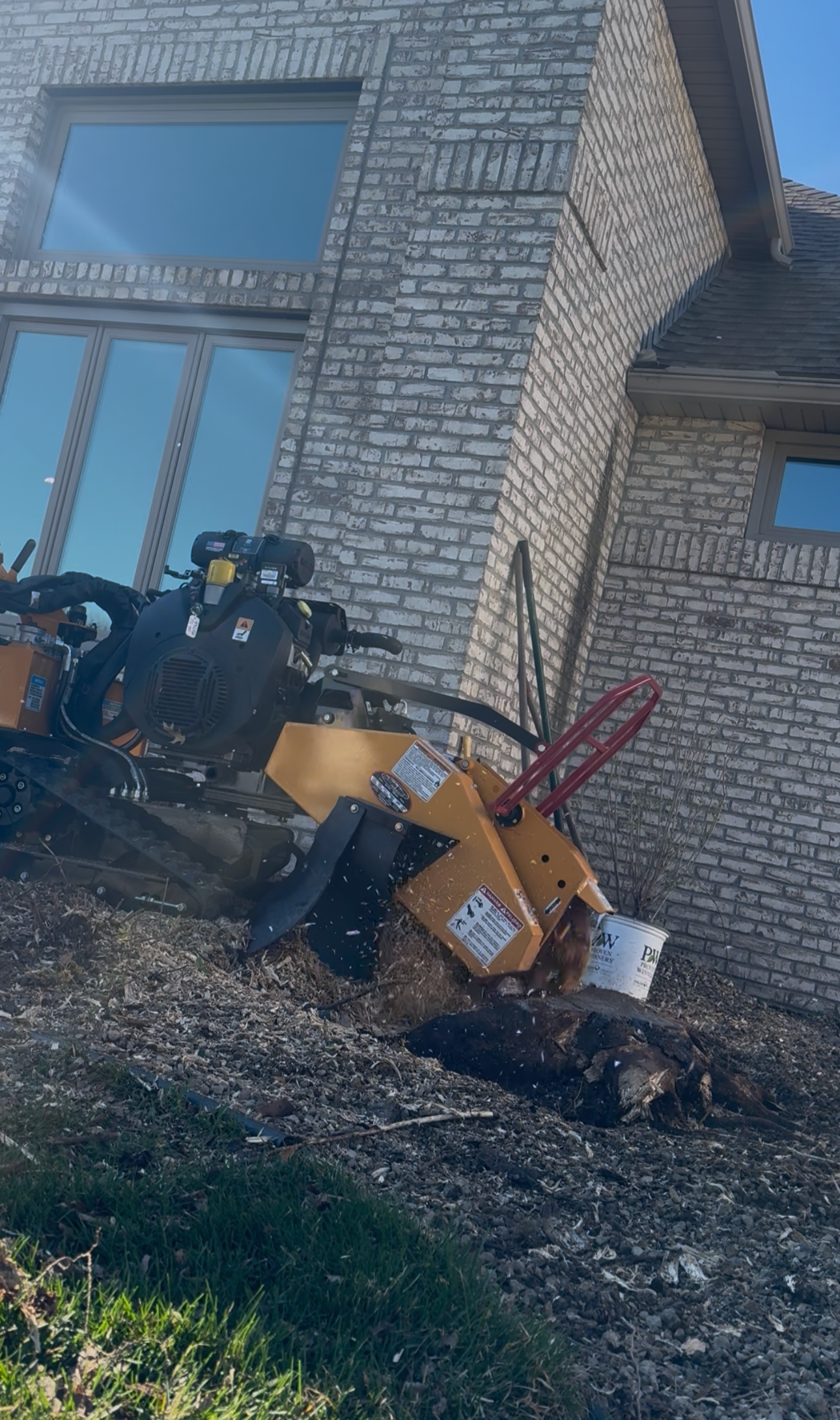 A construction or landscaping machine working on the ground near a brick house with large windows, with dirt and grass in the foreground.