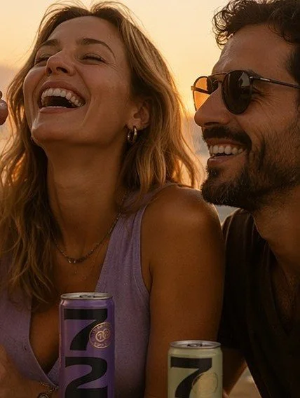 Homem e mulher sorrindo ao lado de latas de bebida, com pôr do sol ao fundo