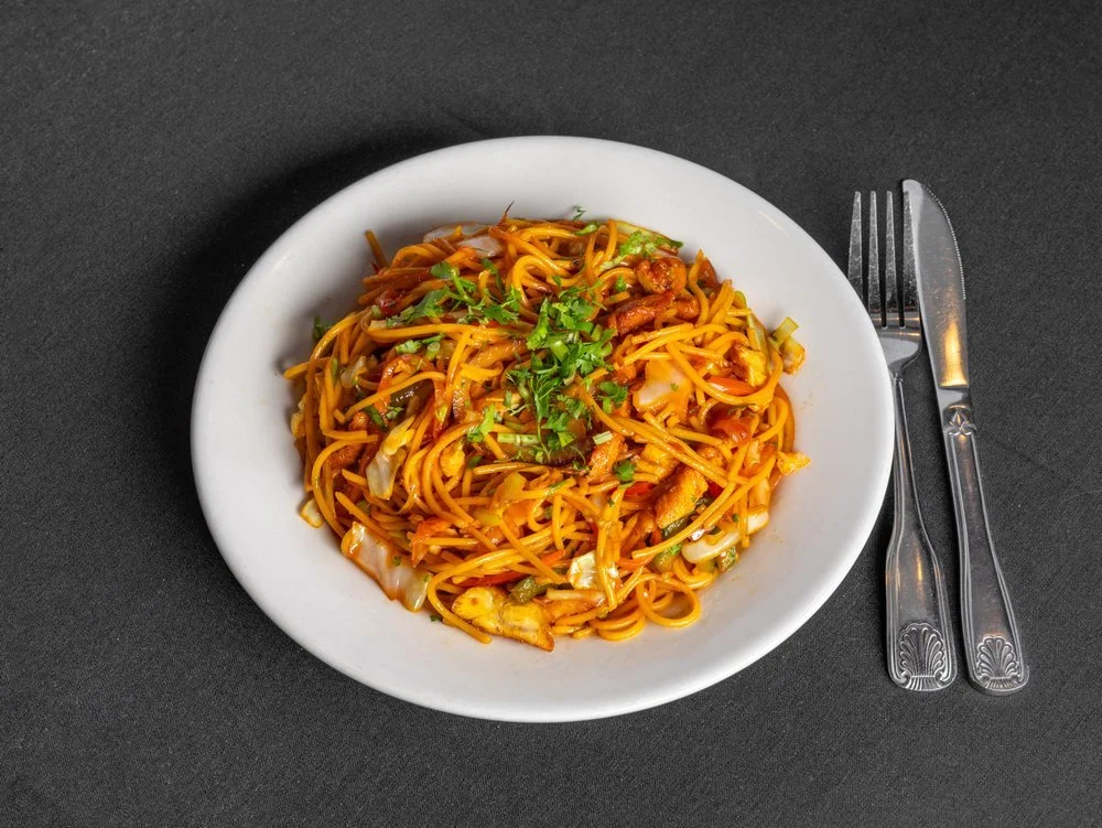 Plate of stir-fried noodles with vegetables and herbs, on a black table with a fork and knife.