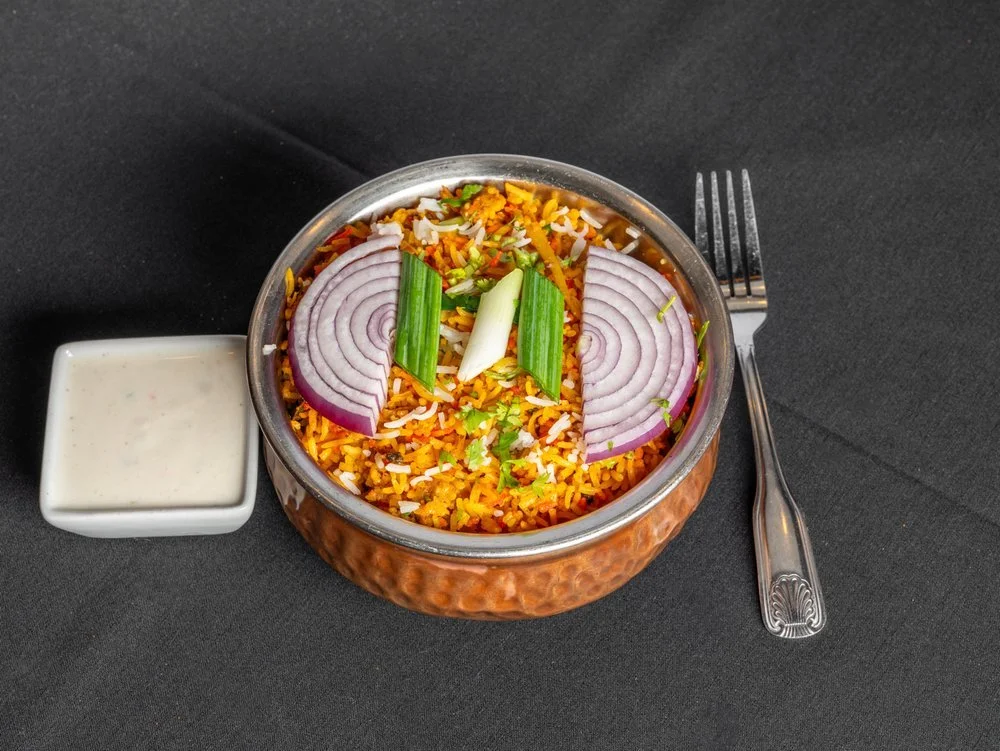 A bowl of Indian vegetable biryani garnished with sliced onions and green onions, served with a side of raita on a black table, with a fork placed beside the bowl.