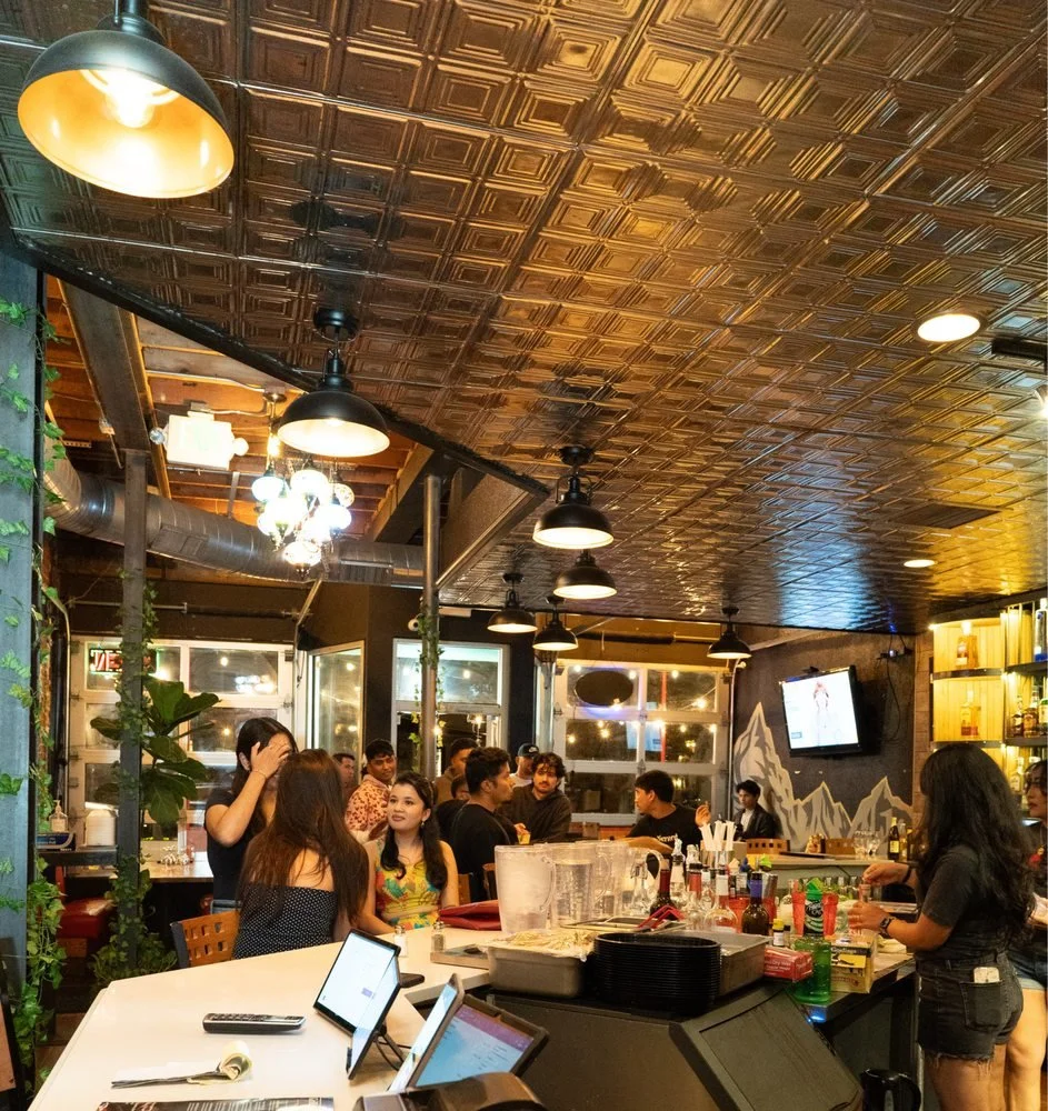 People socializing inside a lively bar or restaurant with warm lighting, wooden decor, a TV screen, and a bar counter with drinks and supplies.