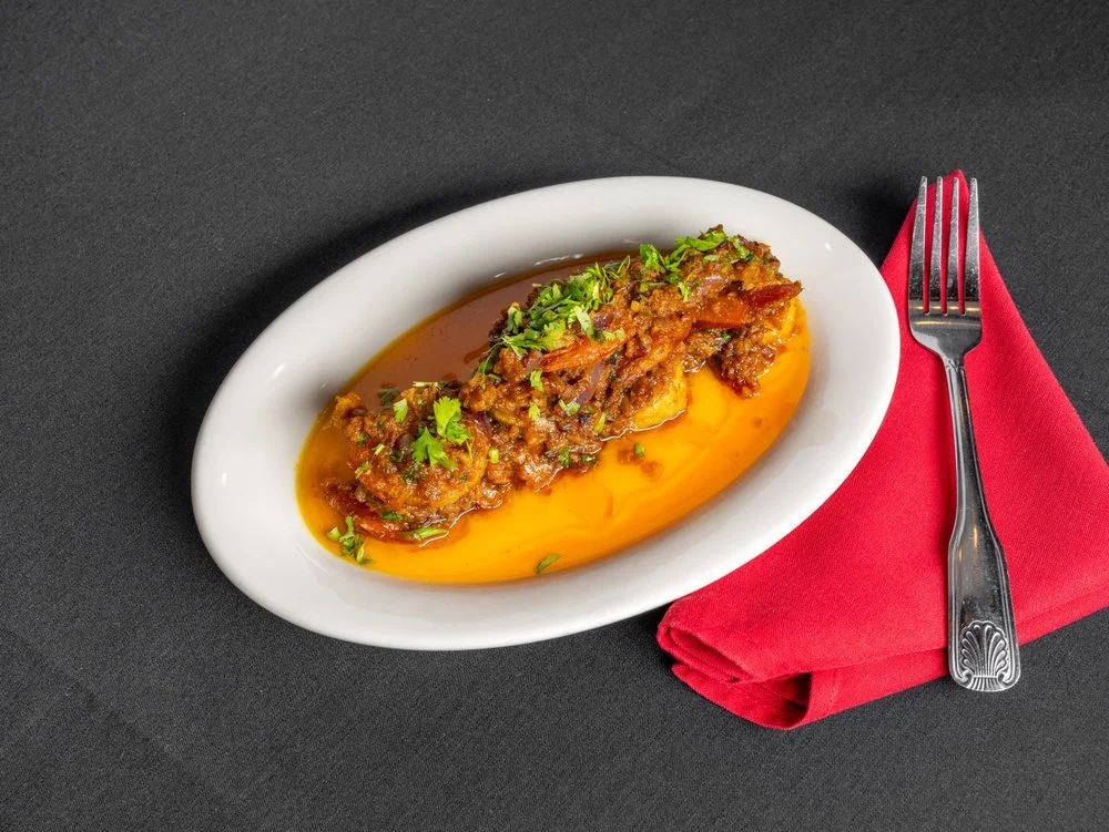 A white oval dish with pulled beef and gravy served on mashed sweet potatoes, garnished with chopped herbs, placed on a dark surface with a red napkin and a fork.