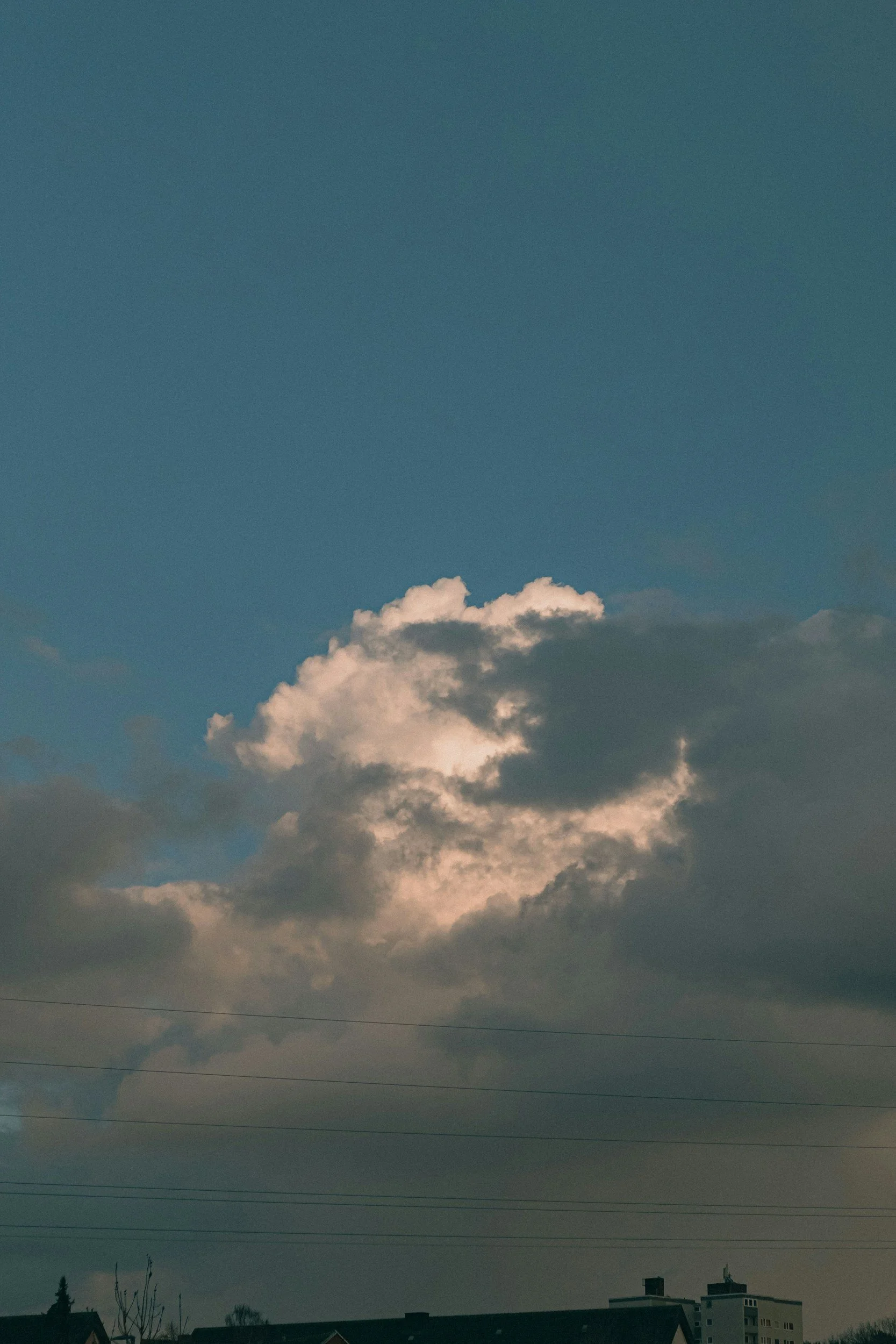 Sky with dark clouds and a few smaller clouds at sunset or sunrise over residential buildings and trees.