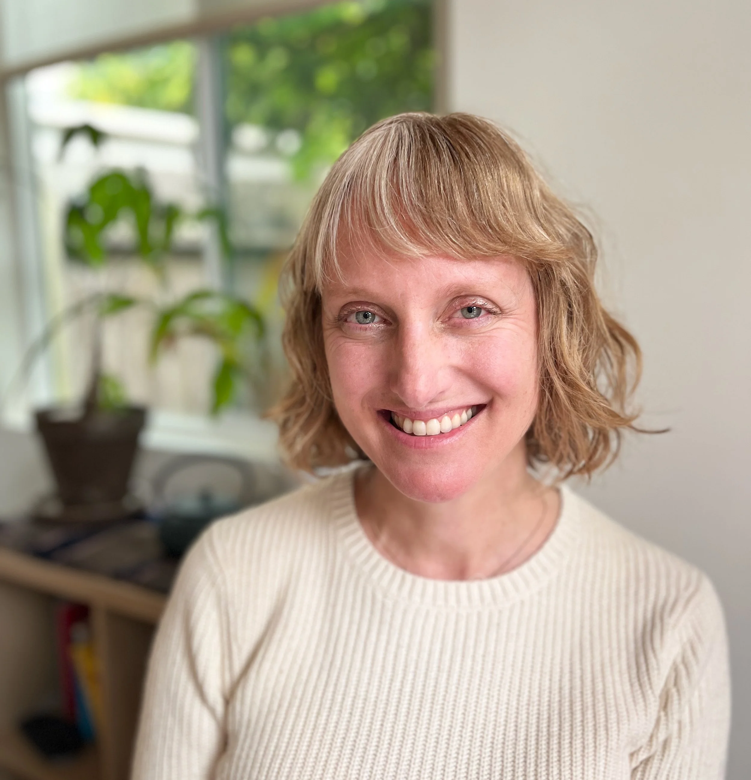 Close-up of a smiling woman with short, light brown hair, wearing a cream-colored ribbed sweater, standing in front of a blurred background with a window and a plant.
