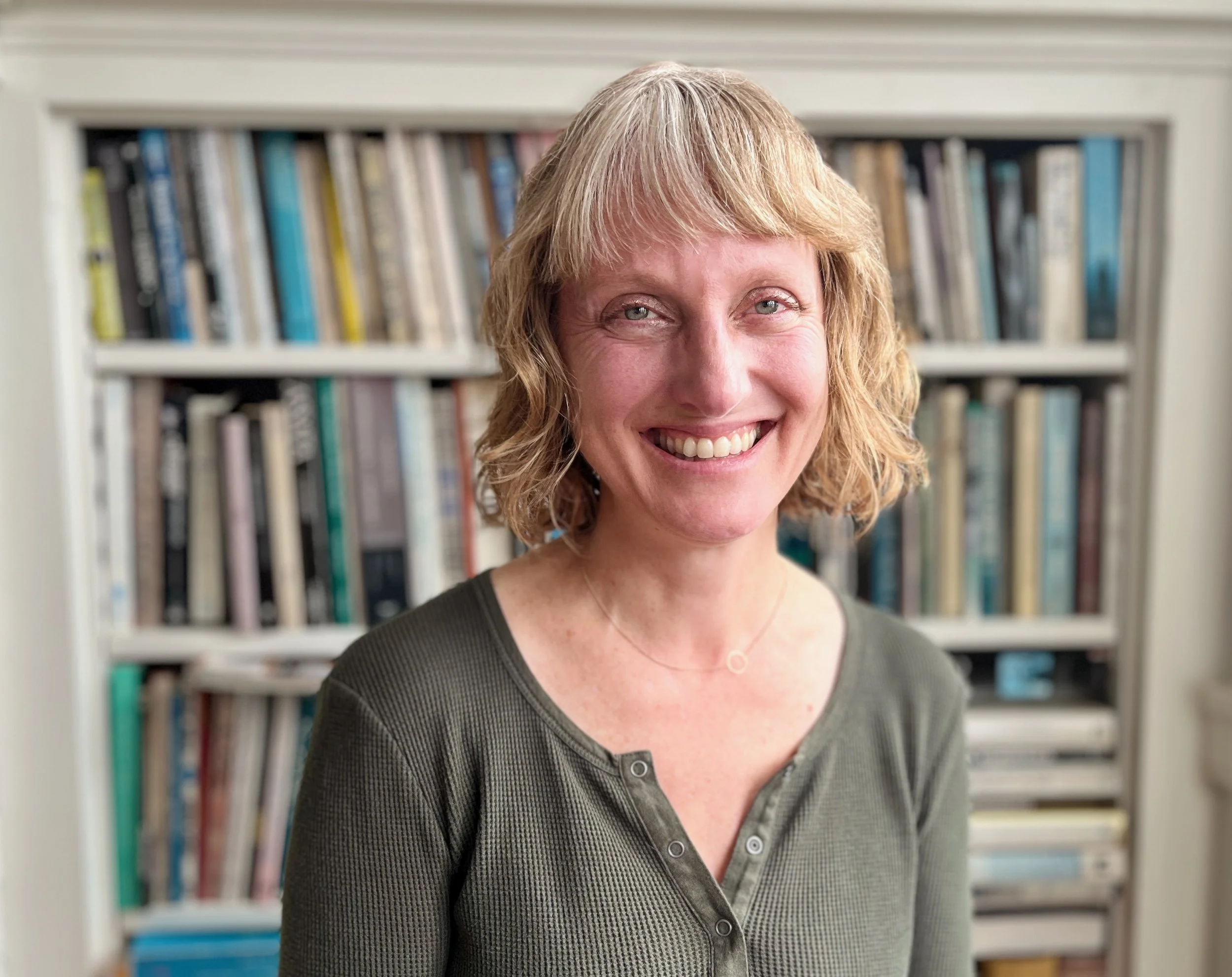 A smiling woman with blonde, wavy hair wearing a dark green top and standing in front of a bookshelf filled with books.