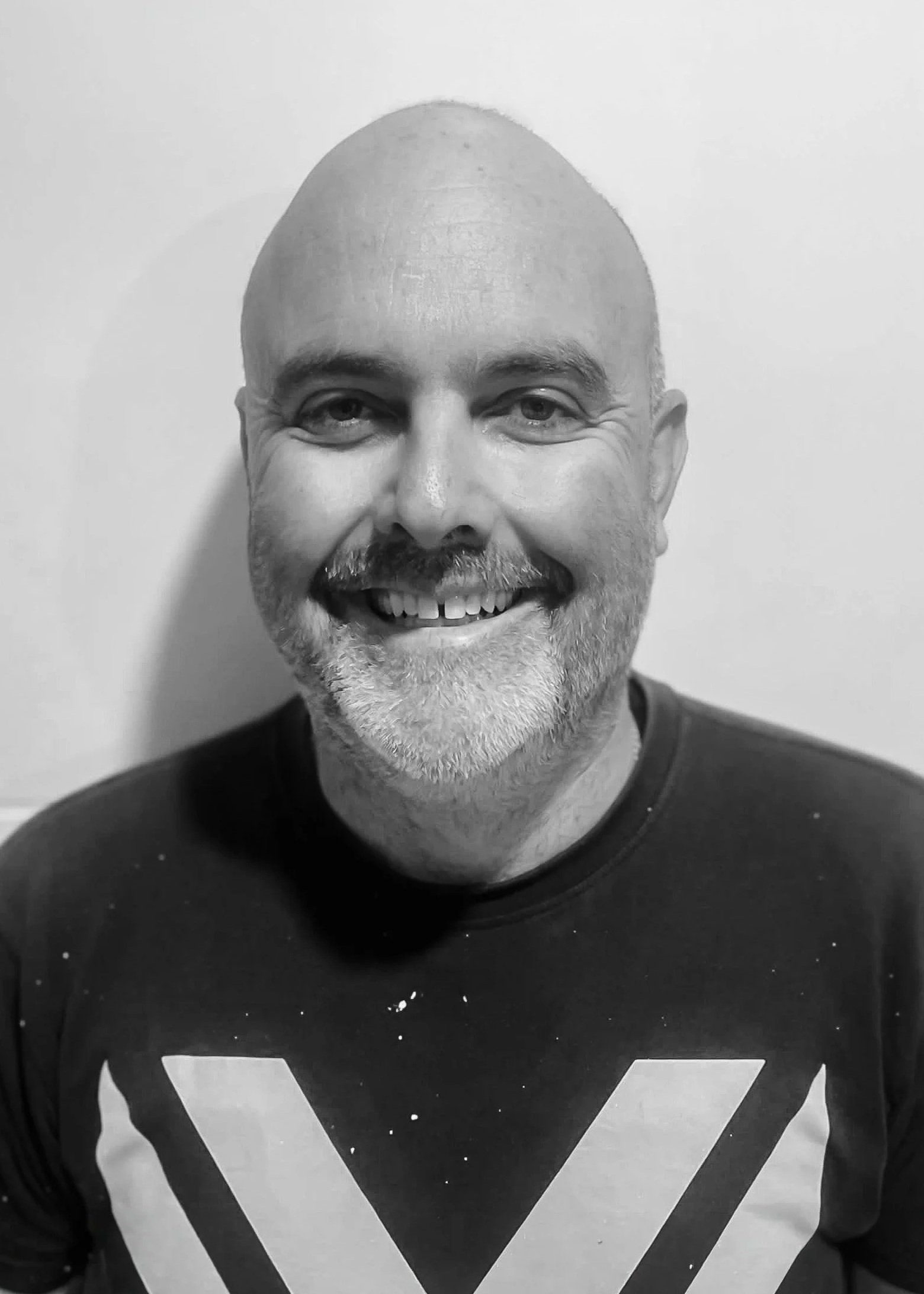 Black and white photo of a man with a beard, smiling, wearing a shirt with a large geometric 'V' on the front, standing against a plain wall background.