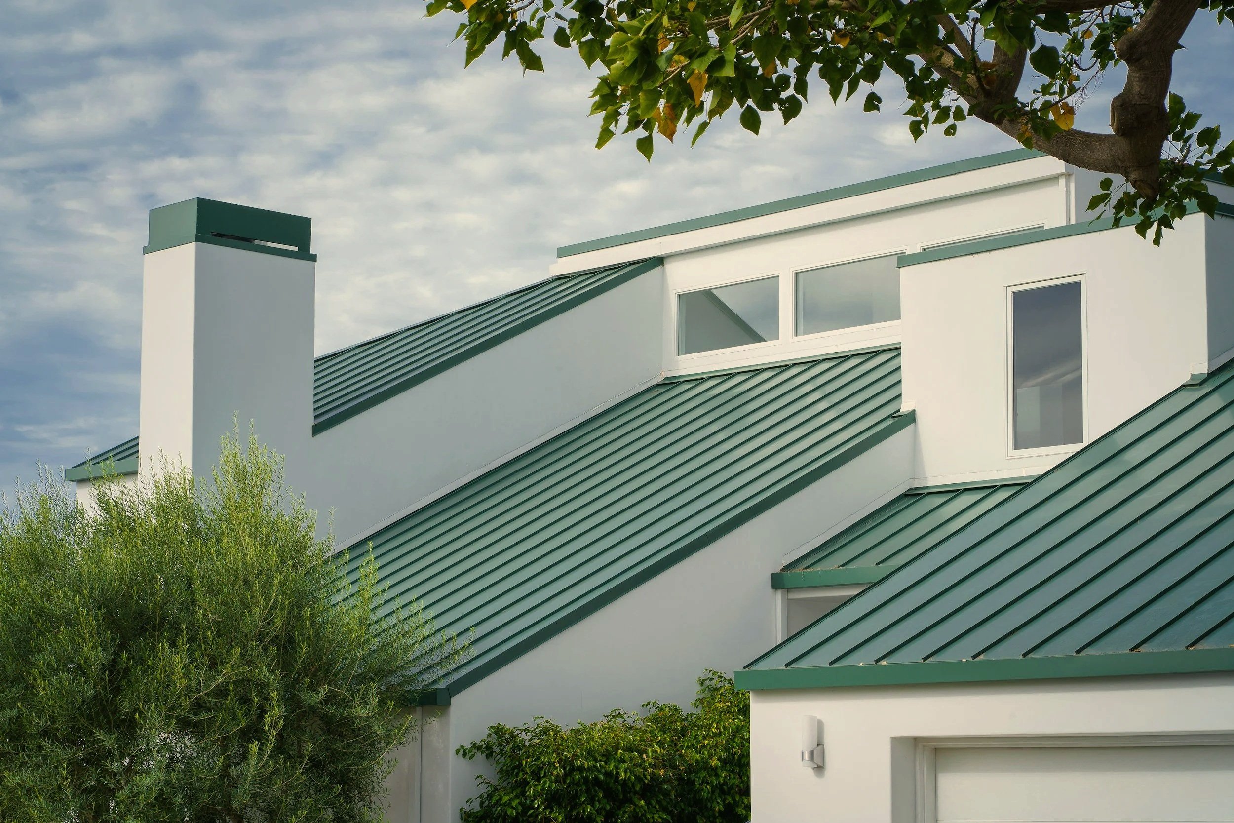 A modern house with a sloped green metal roof, white walls, and a large window, partially obscured by trees and foliage, under a partly cloudy sky.