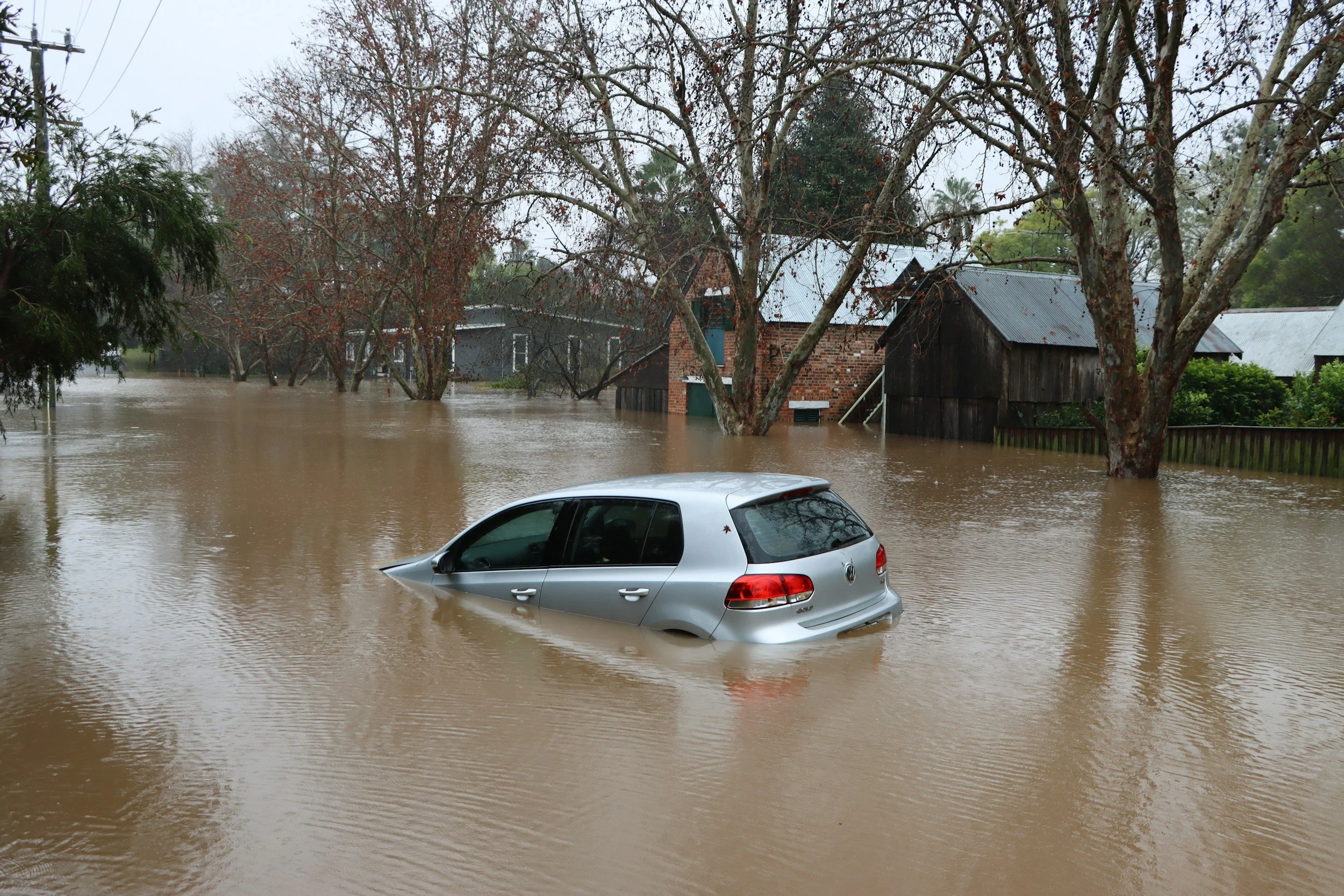 A flooded street with a silver car partially submerged in brown water, nearby houses, and trees with some leaves, indicating floodwaters after heavy rain.
