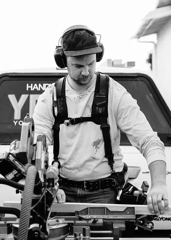 A young man working with a power saw outdoors, wearing earmuffs, a cap, a workbelt, and a white hoodie with paint stains, standing in front of a vehicle.